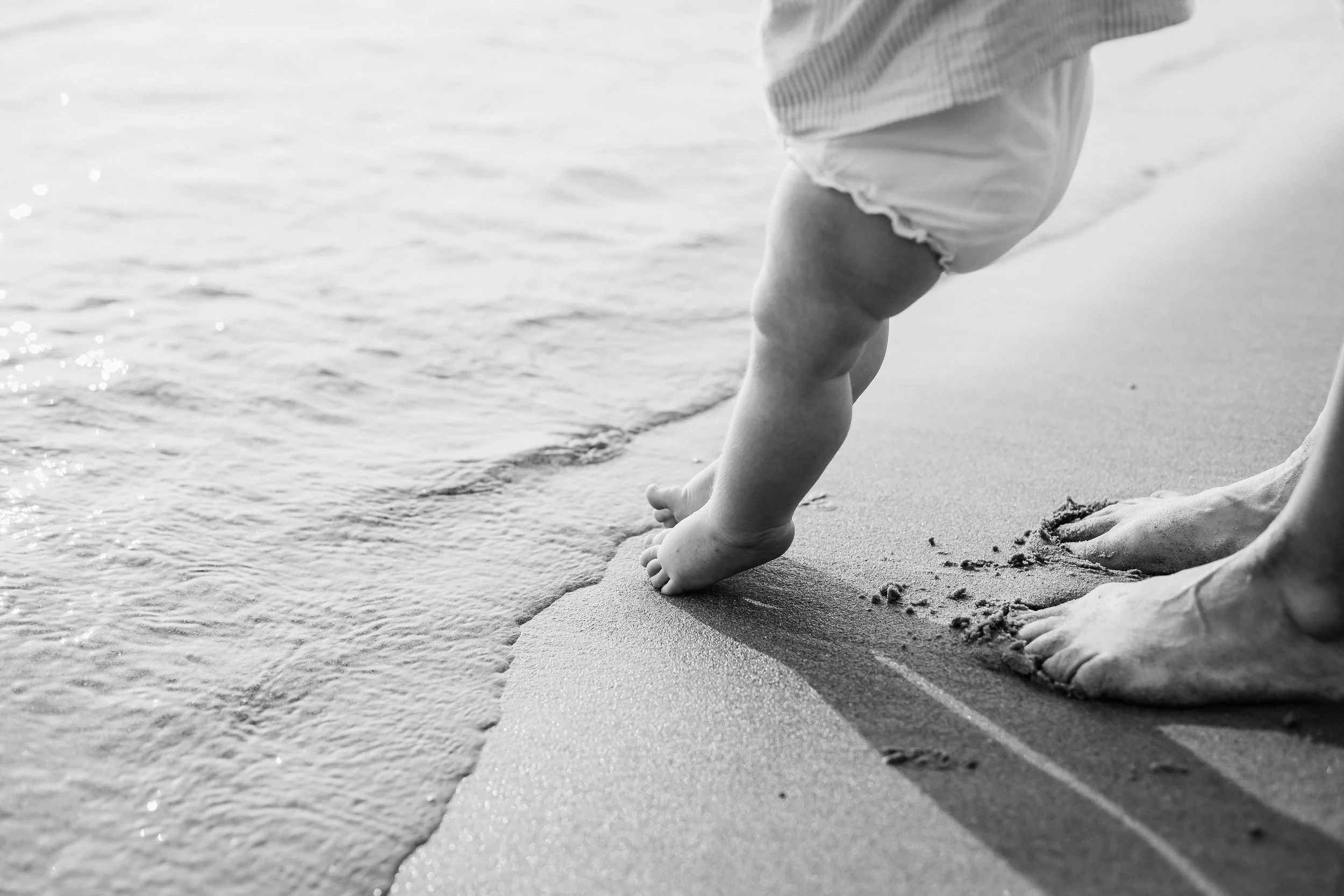 A child learning to walk on the sandy beach, supported by an adult's feet, near the shoreline. Lifestyle newborn session shot in Evanston IL. Grosse Point Lighthouse Evanston