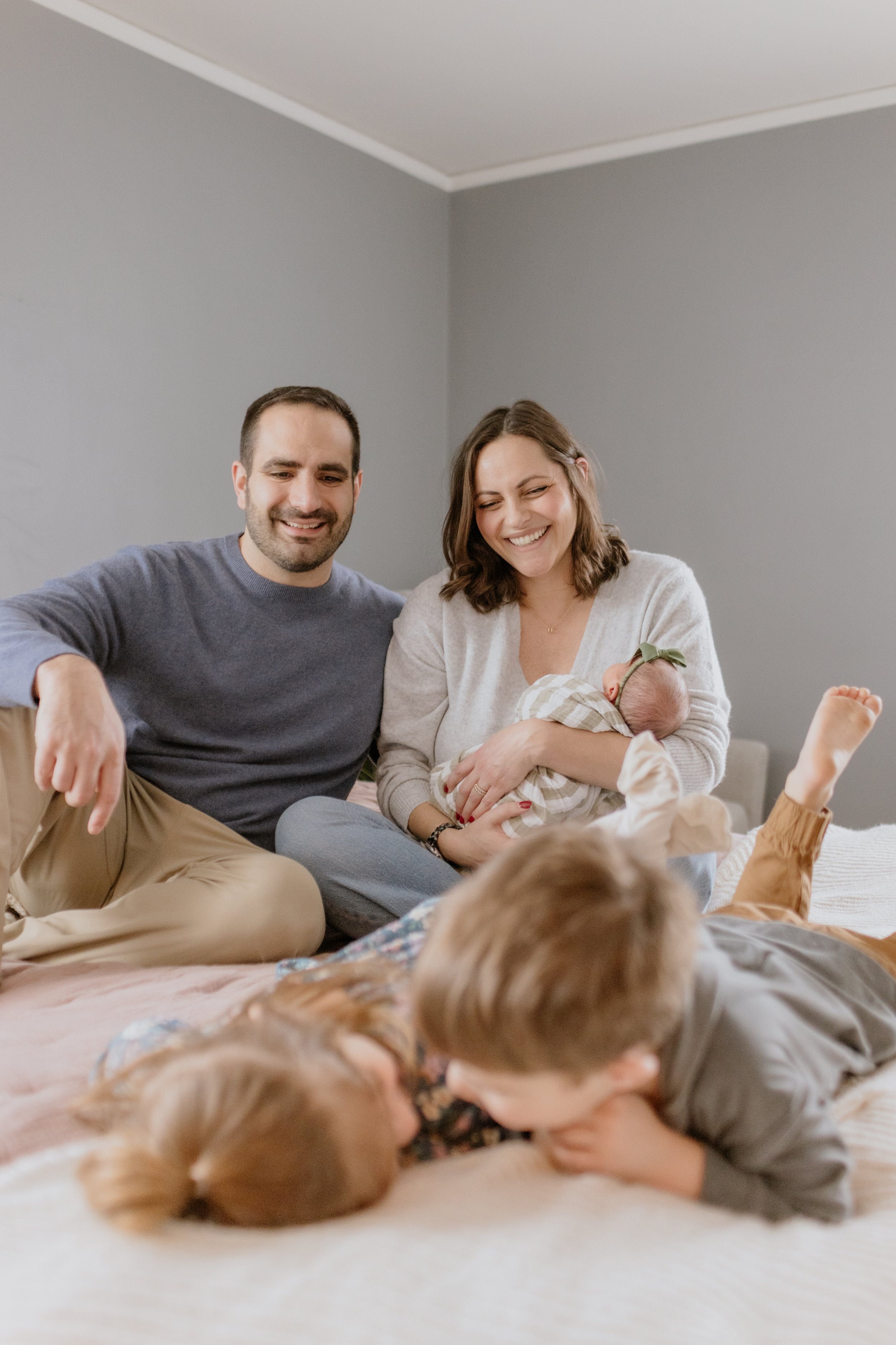 A family of four, including two children, a mother holding a newborn baby with a green headband, and a father, sitting on a bed in a bedroom with gray walls. The children are lying on the bed, and the mother and father are smiling. Lifestyle Newborn 