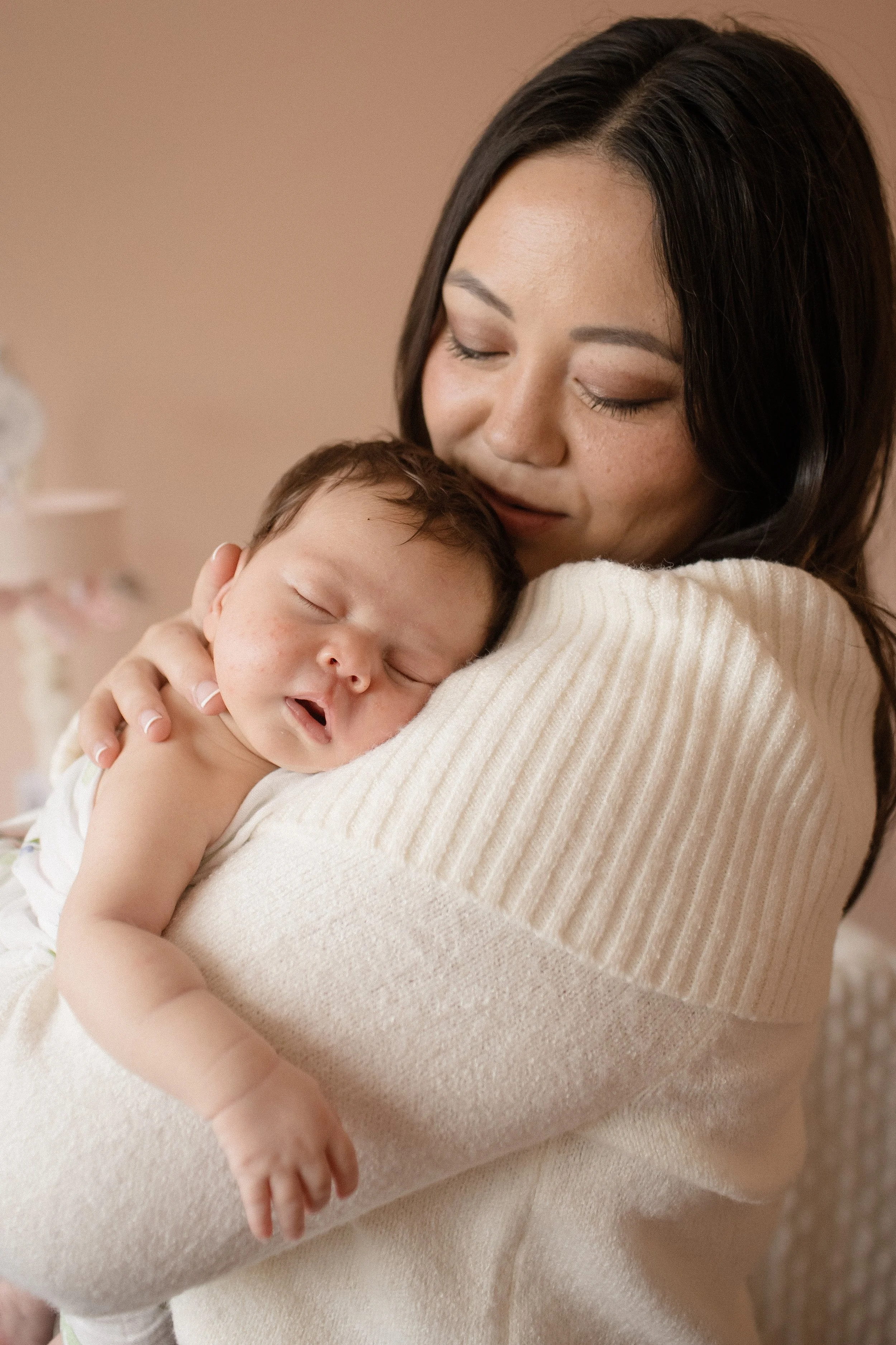 A woman is holding a sleeping baby close to her chest, both with a warm tender expression. Lifestyle newborn session shot in Chicago IL.