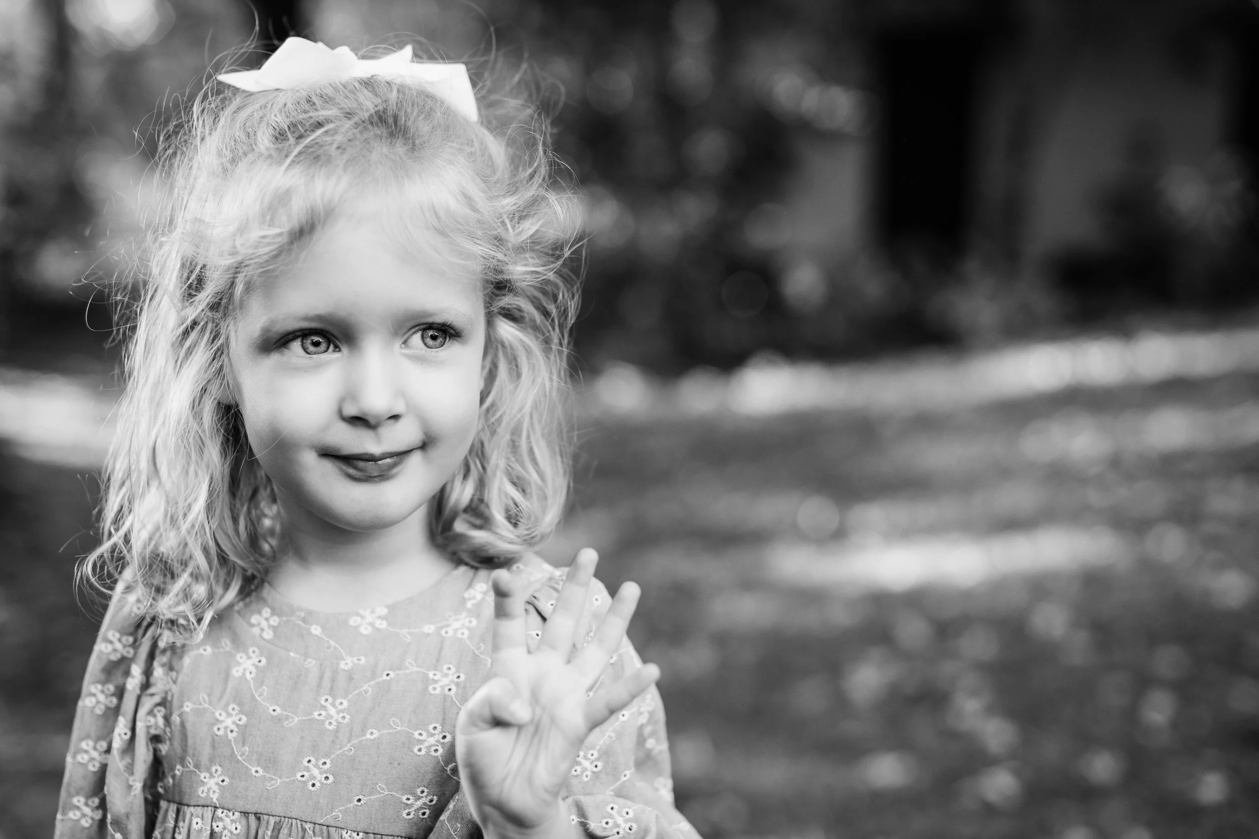 A young girl with curly hair and a bow on her head, making a peace sign with her hand and smiling, outdoors in black and white.