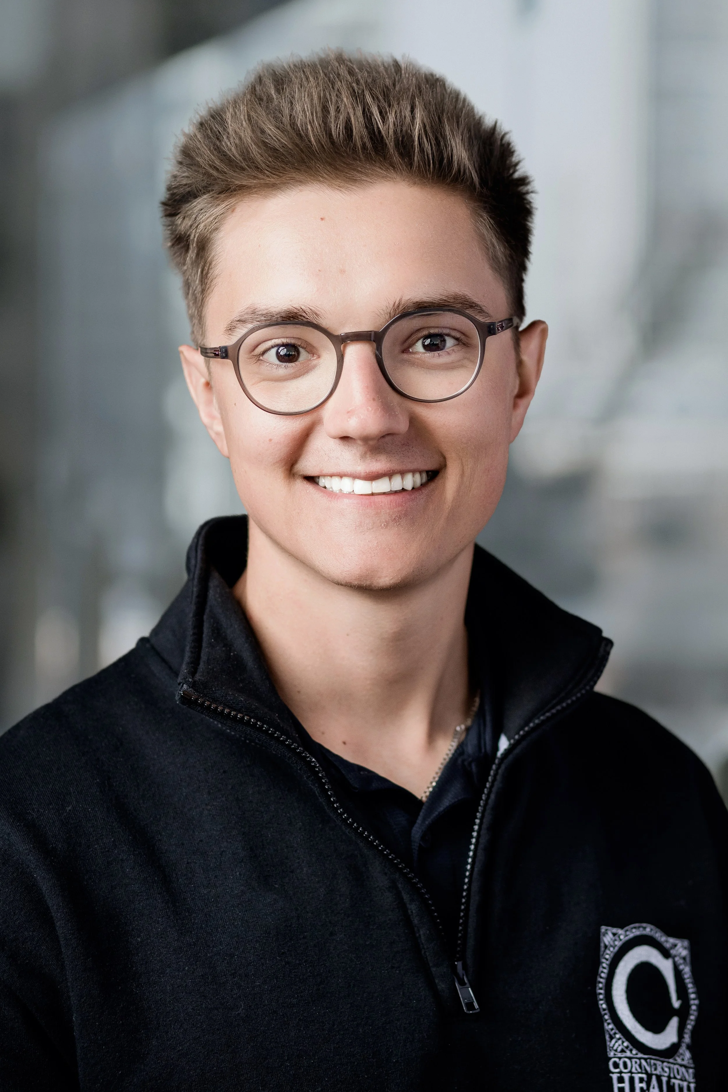 A young man with short brown hair, glasses, and a bright smile wearing a black zip-up jacket with a logo on the chest that reads 'Cornerstone Health'. Headshot photography session in Evanston, IL.