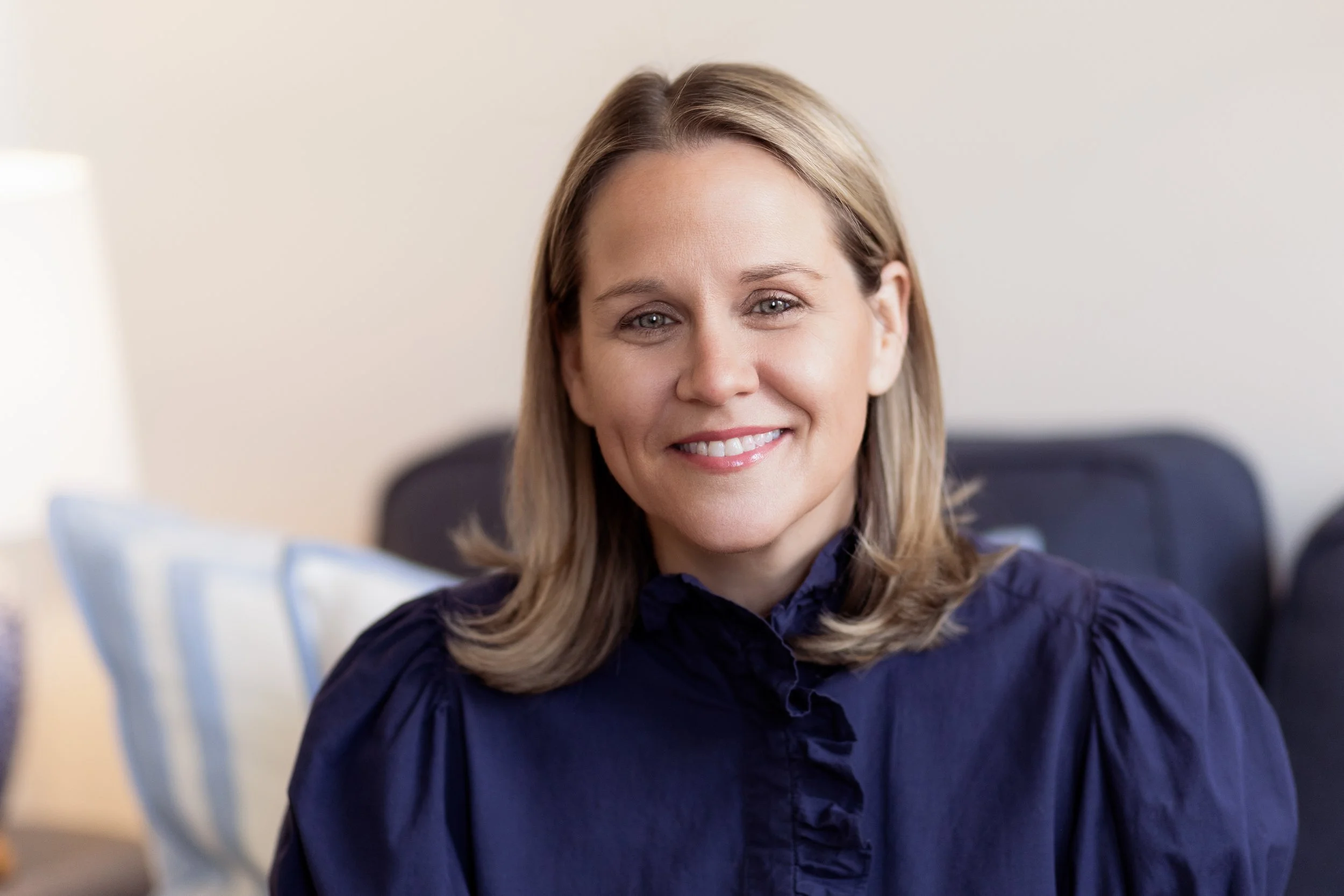A woman with shoulder-length blonde hair wearing a navy blue blouse, smiling while sitting on a couch in a well-lit room with white walls and patterned pillows.