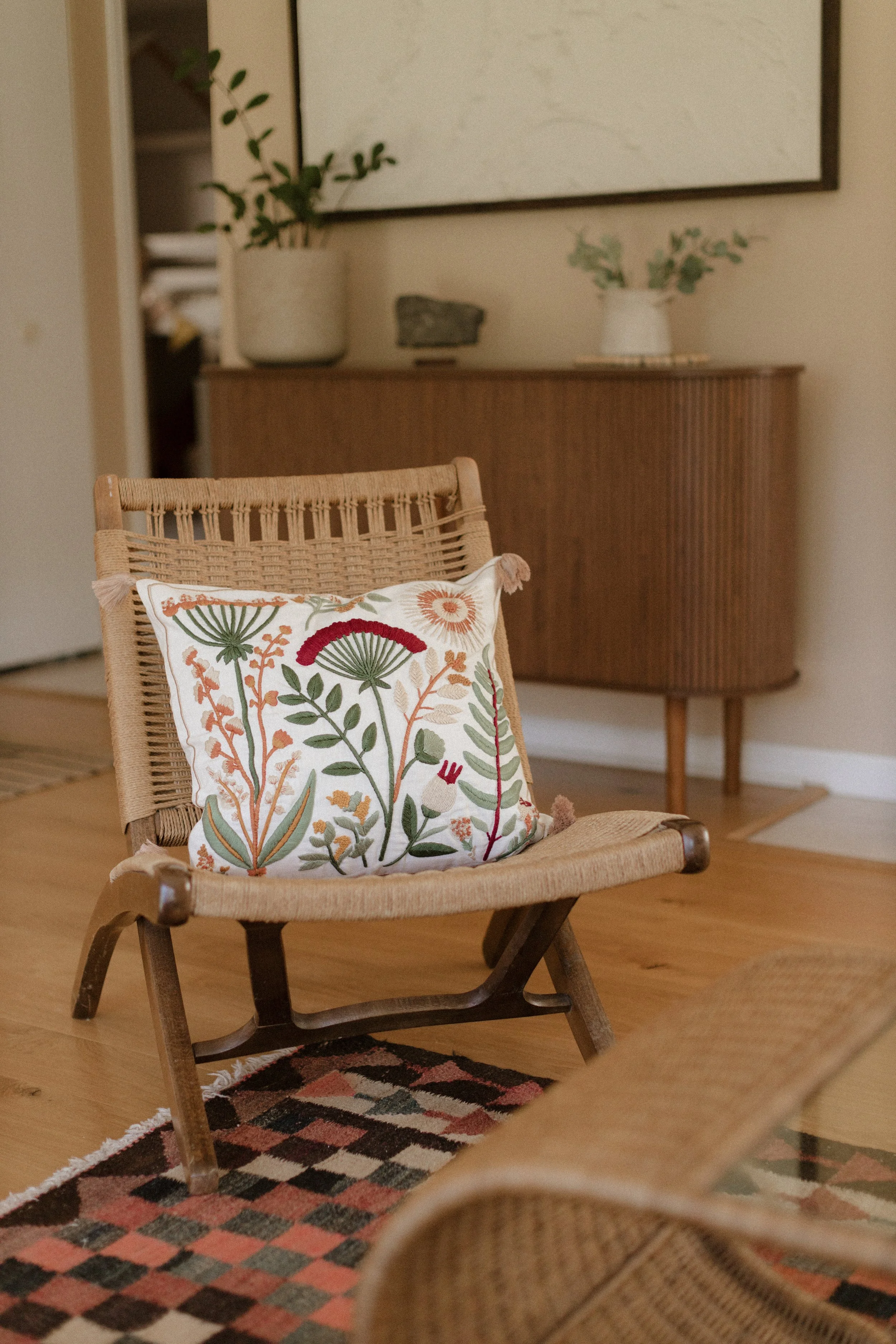 A wicker chair with a decorative pillow featuring a colorful floral embroidery, placed in a cozy room with a wooden sideboard in the background, and a patterned rug on the wooden floor. Product Photography Shoot Evanston, IL.