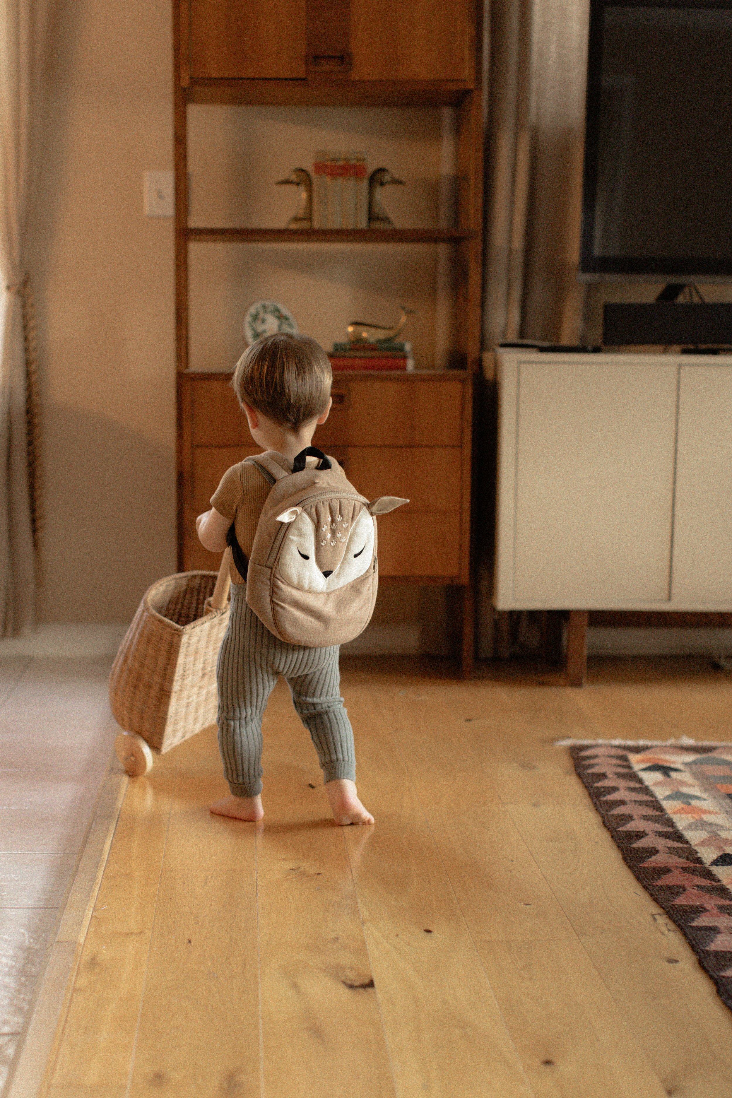 A young child with a beige backpack featuring a cute animal face, pulling a small wicker cart, standing in a cozy living room with wooden floors, shelves, and a television. Product Photography Shoot Evanston, IL.