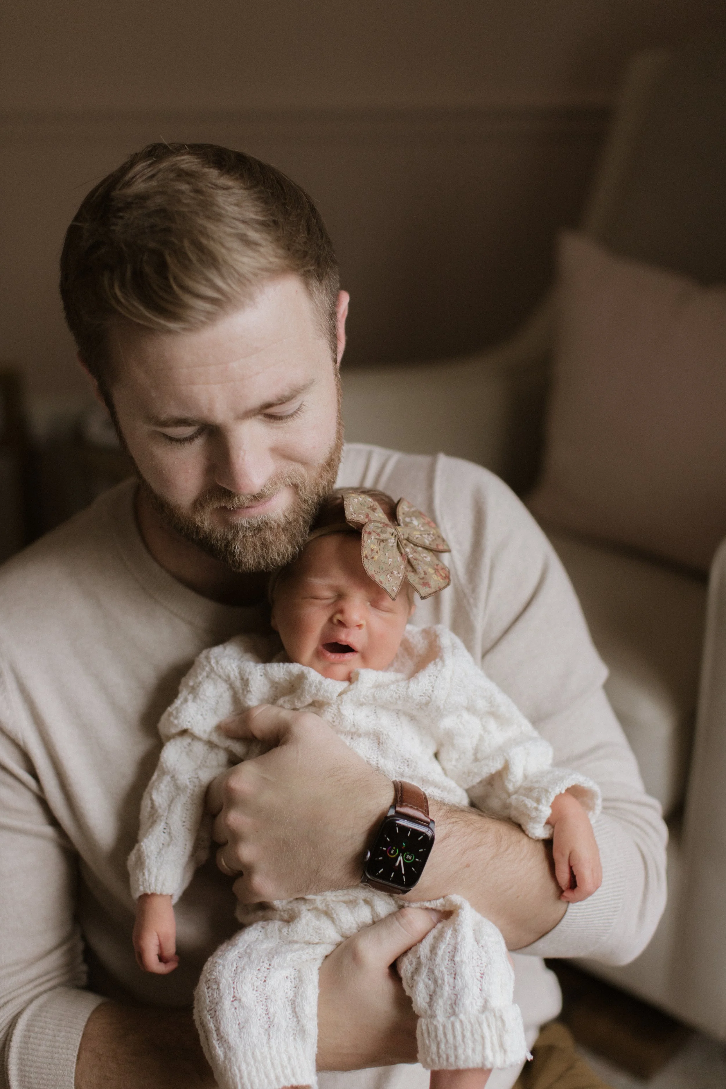 A man with a beard holding a crying baby girl wearing a large bow headband and a cream knitted outfit in a cozy room.