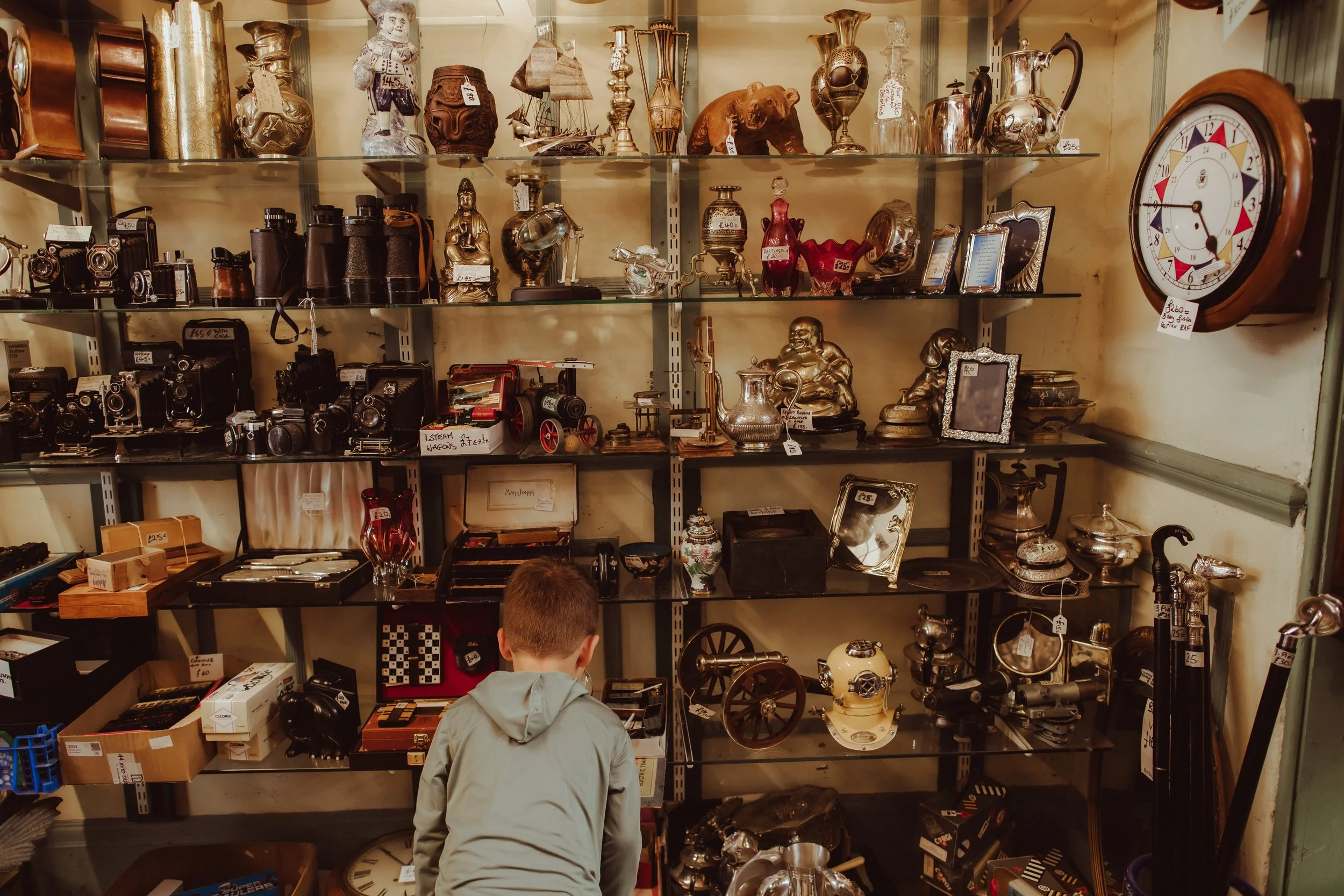 A young boy shopping at an antique store with shelves filled with vintage items including cameras, clocks, vases, figurines, and decorative objects.