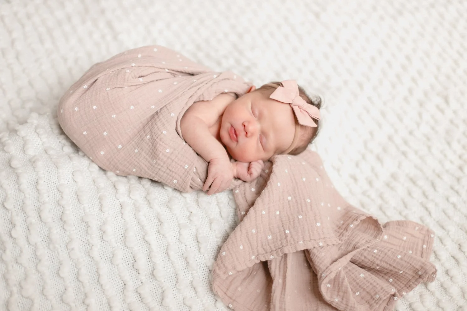Newborn baby girl sleeping wrapped in a pink, dotted blanket with a matching pink bow headband on a textured white surface. Lifestyle newborn session shot in Evanston IL.