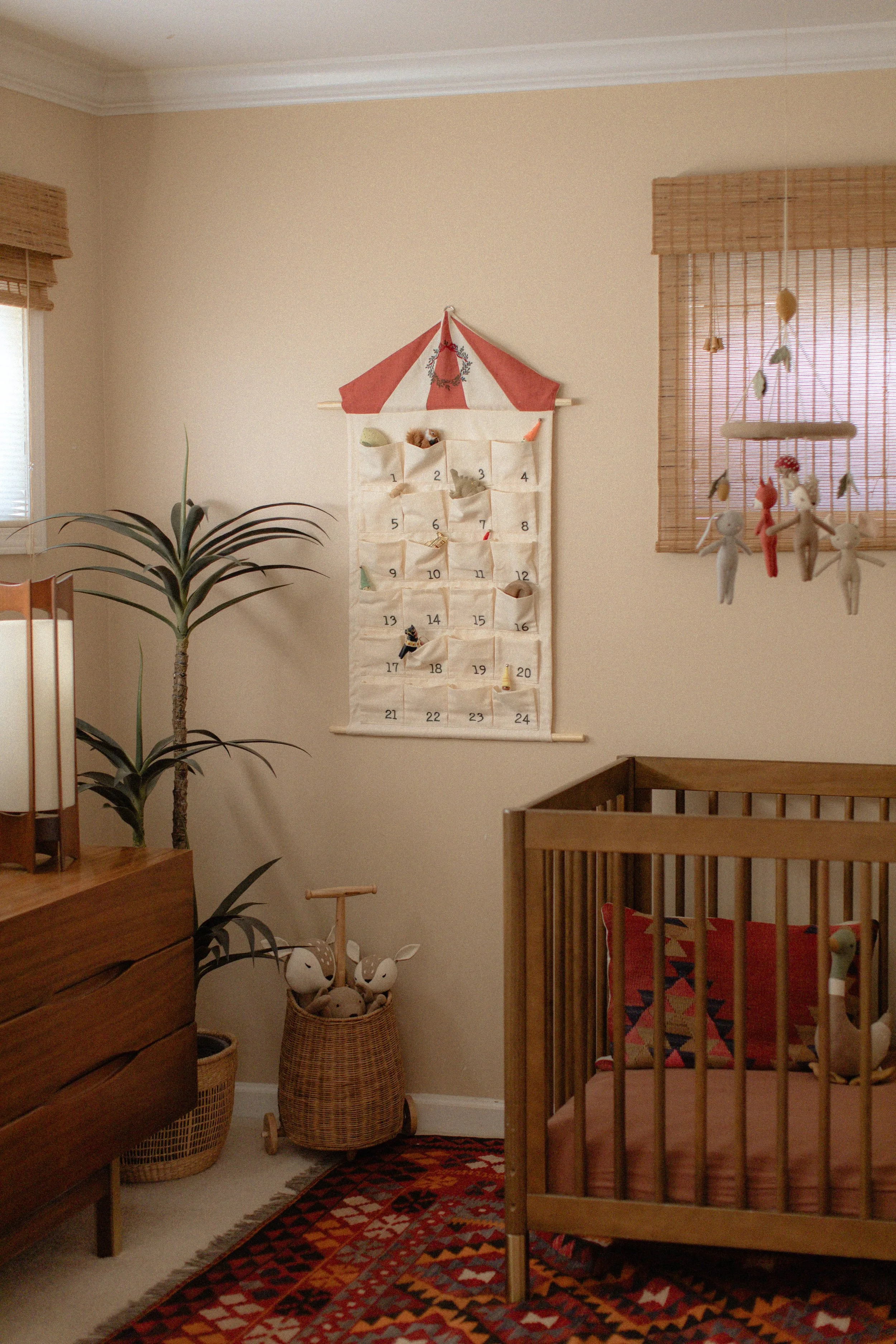 A cozy nursery room with a wooden crib, a tall plant, a woven basket with plush toys, a patterned rug, and a wall hanging with a calendar and small plush animals. Product Photography Shoot Evanston, IL.