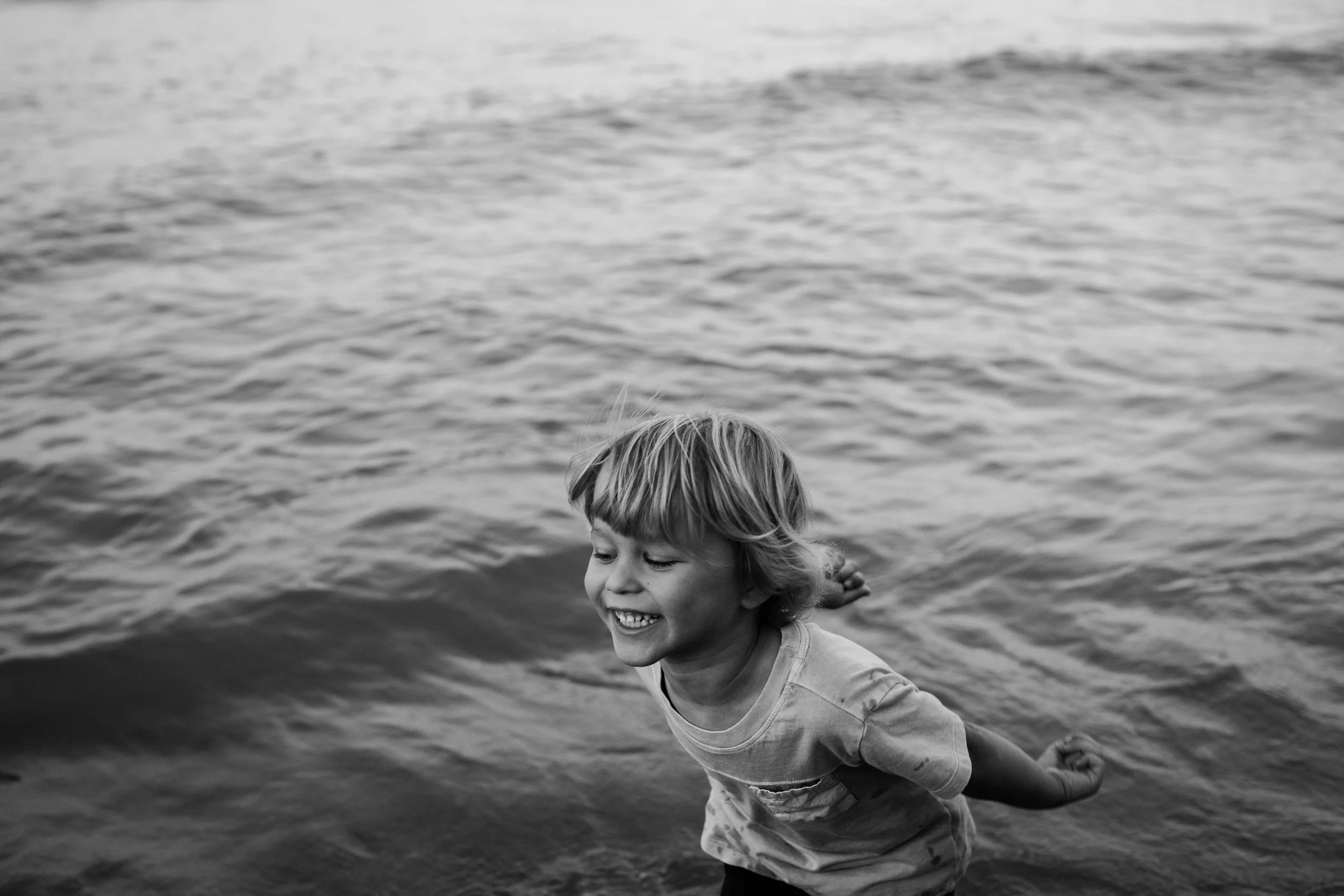 A young boy with a big smile running out of the water at the beach in black and white.