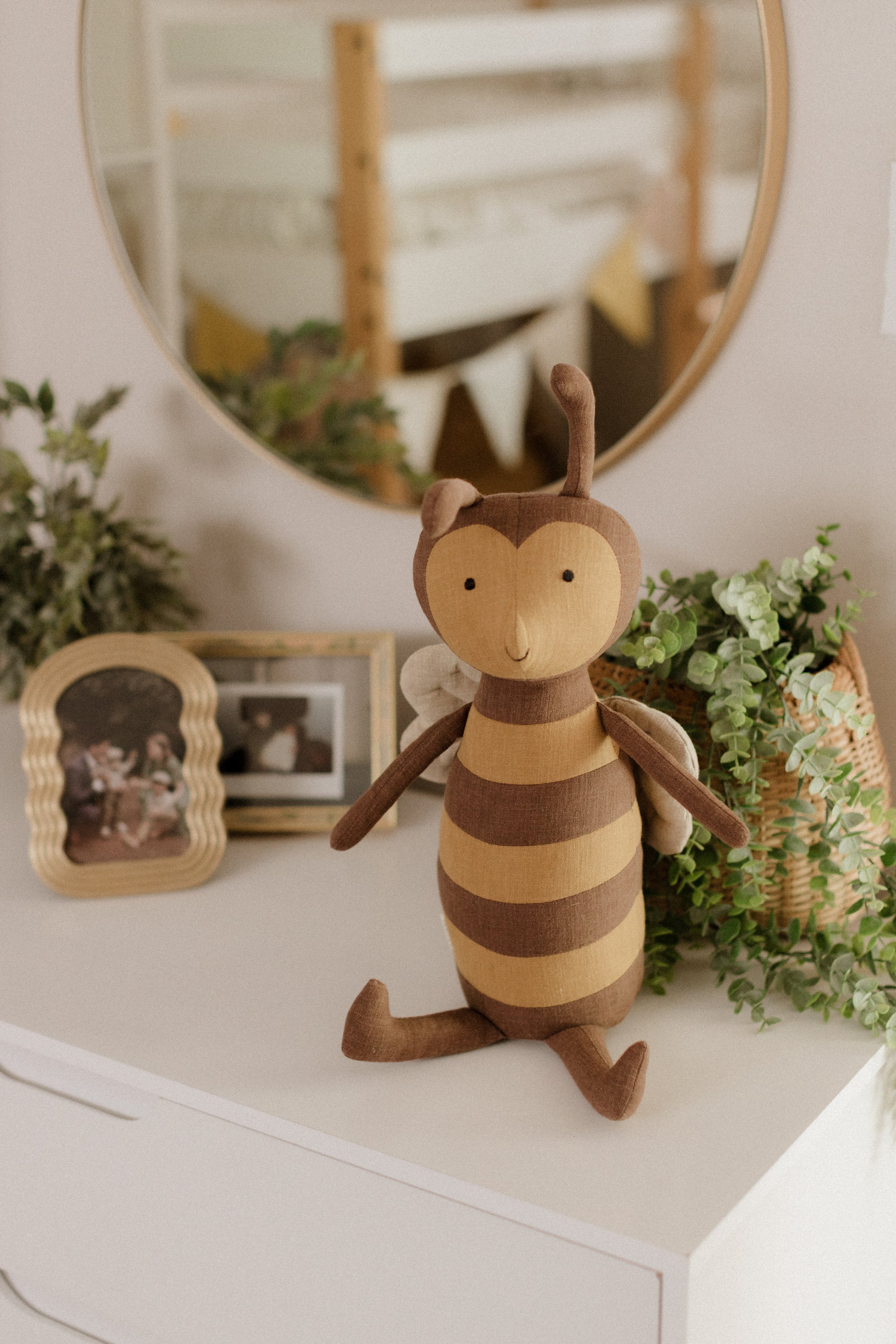 A plush toy of a bee with a smiling face, sitting on a white dresser next to framed photos and green plants, with a round mirror reflecting a nursery in the background.
