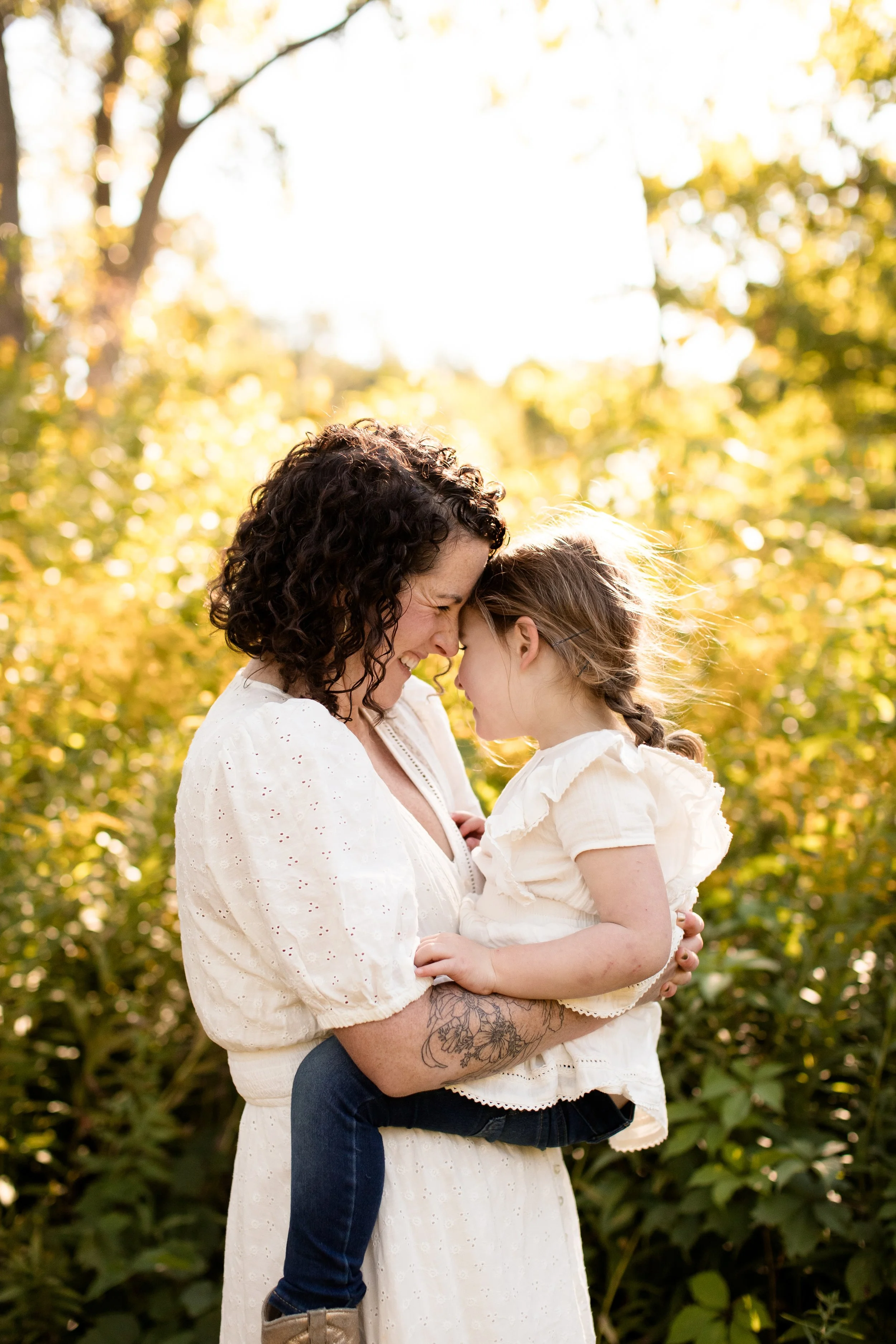 A woman with curly dark hair and a tattoo holds a young girl with braided hair outdoors during golden hour. They touch foreheads, smiling softly, with trees and sunlight in the background.