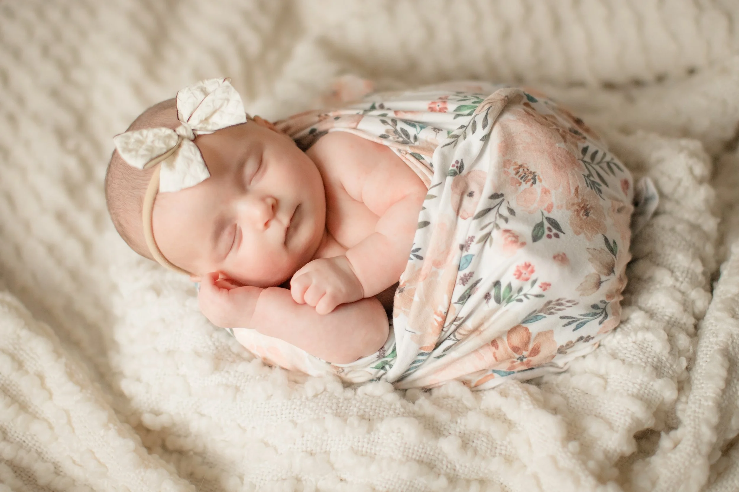 A sleeping baby girl with a floral headband, wrapped in a floral blanket, lying on a soft, cream-colored textured blanket.