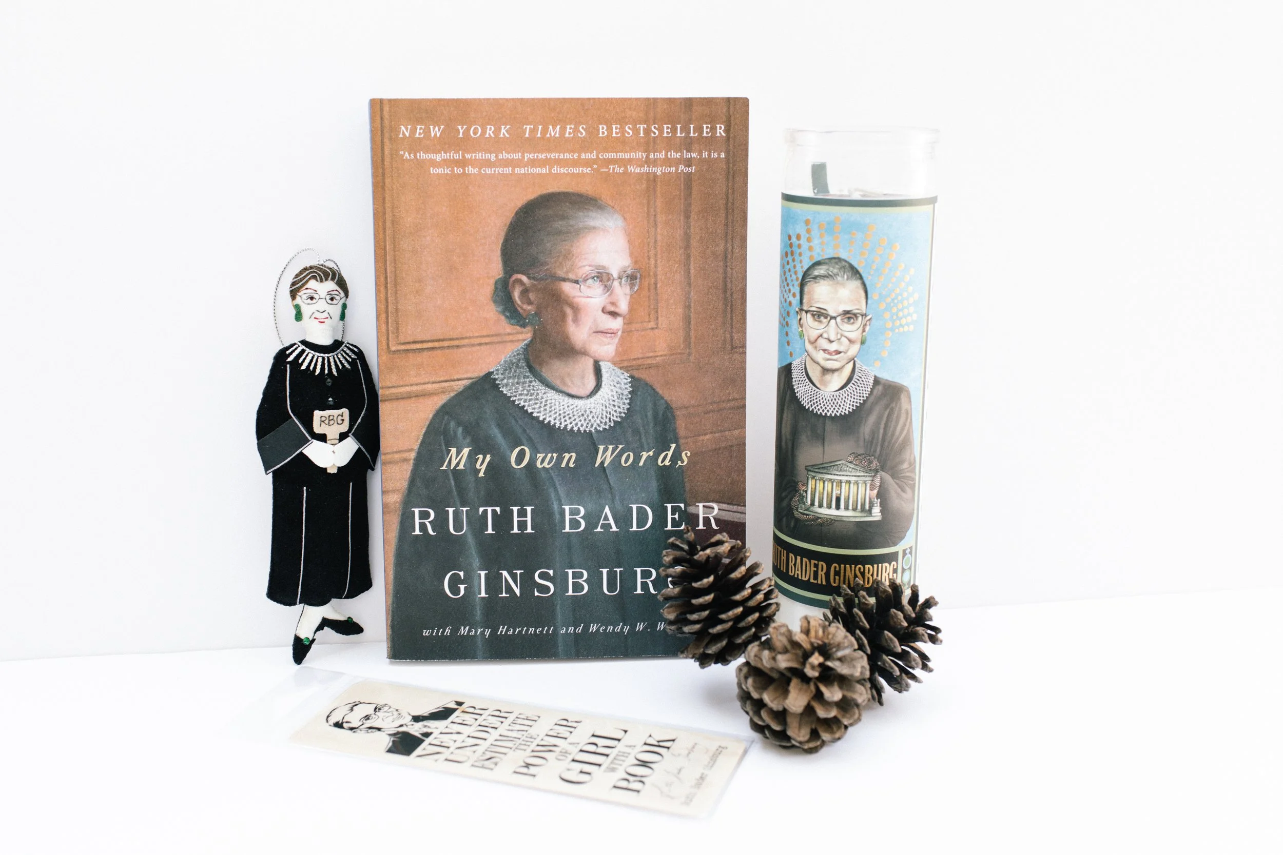 A collection of Ruth Bader Ginsburg-themed items, including a book titled 'My Own Words' by Ruth Bader Ginsburg, a tumbler with her portrait, a paper doll of Ruth Bader Ginsburg, and two pinecones. Product Photography Shoot Evanston, IL.