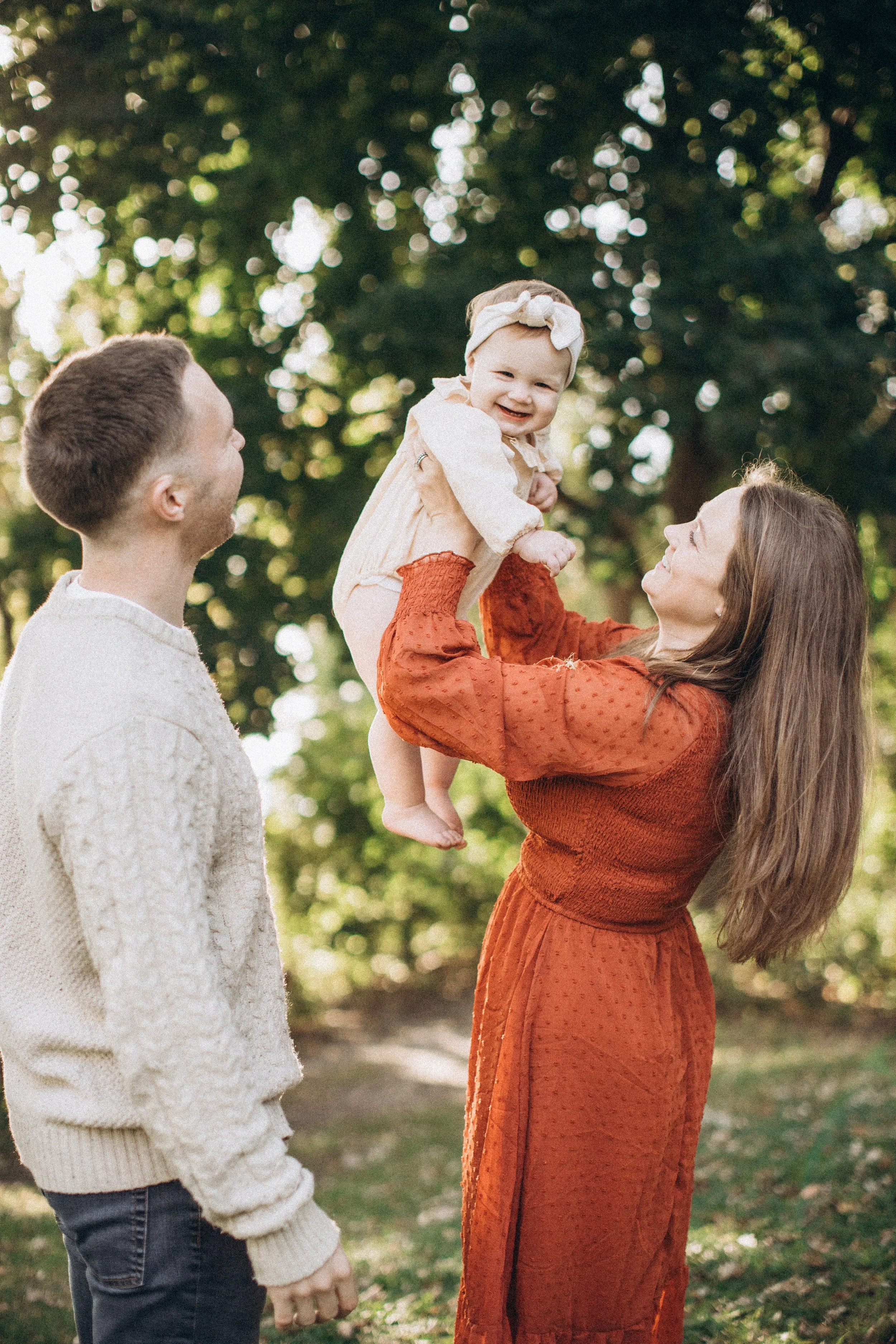 A woman lifting a smiling baby girl with a bow in her hair, while a man watches outdoors during daytime.