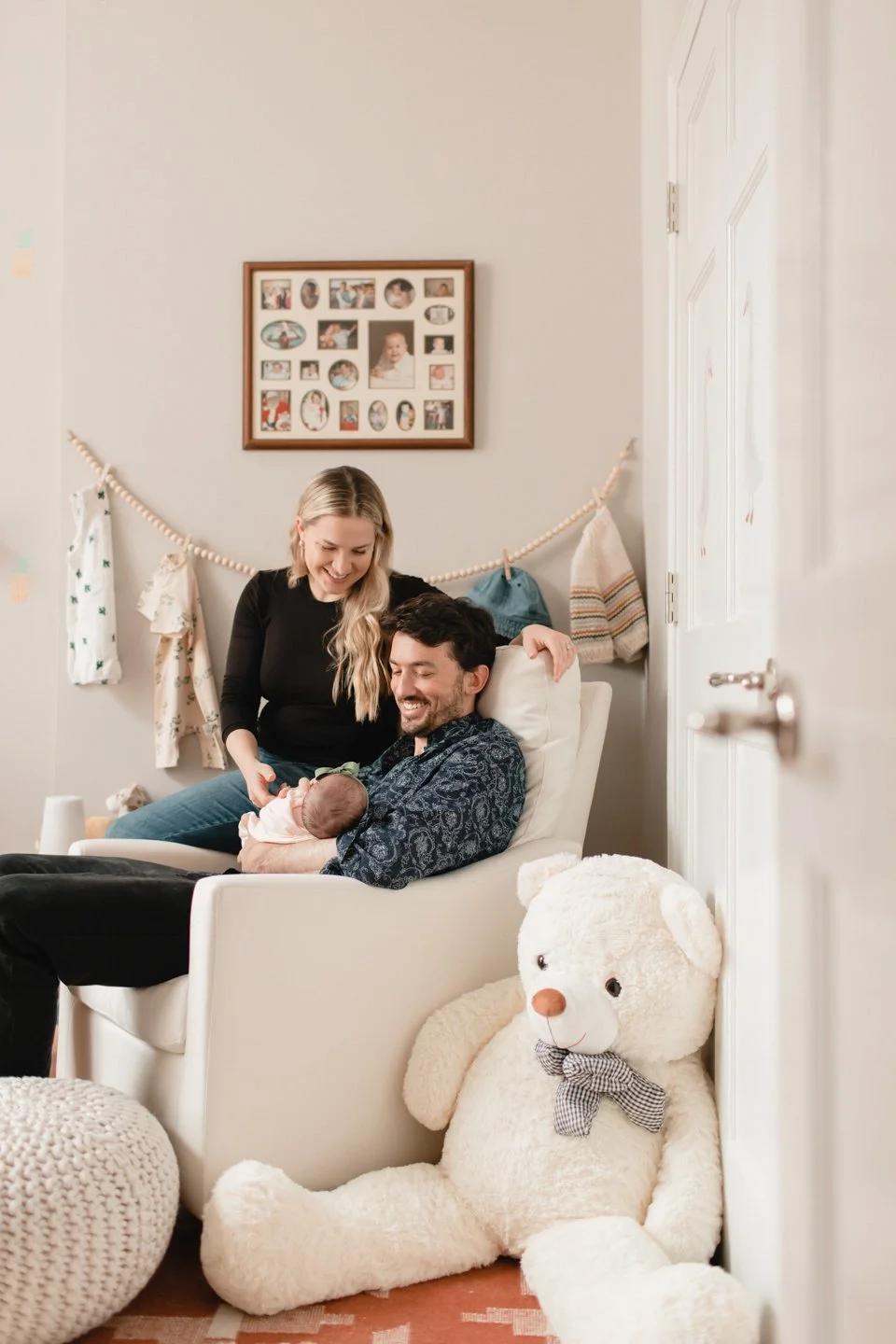 A happy family scene with a smiling man holding a newborn baby in a living room, a woman leaning over and touching the baby, and a large teddy bear on the floor.