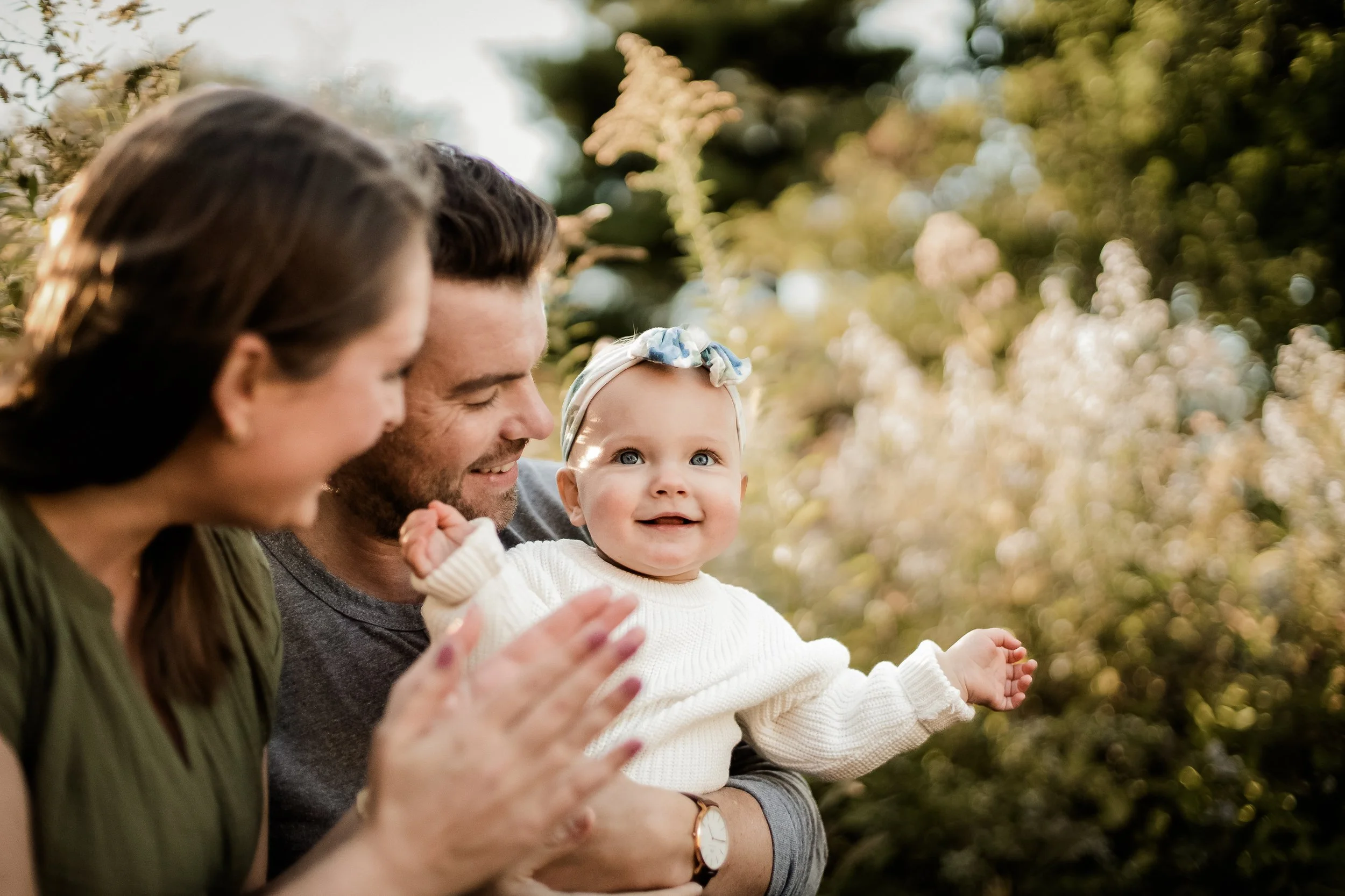 A happy family consisting of a woman, a man, and a baby girl outdoors among flowering bushes, enjoying a sunny day.