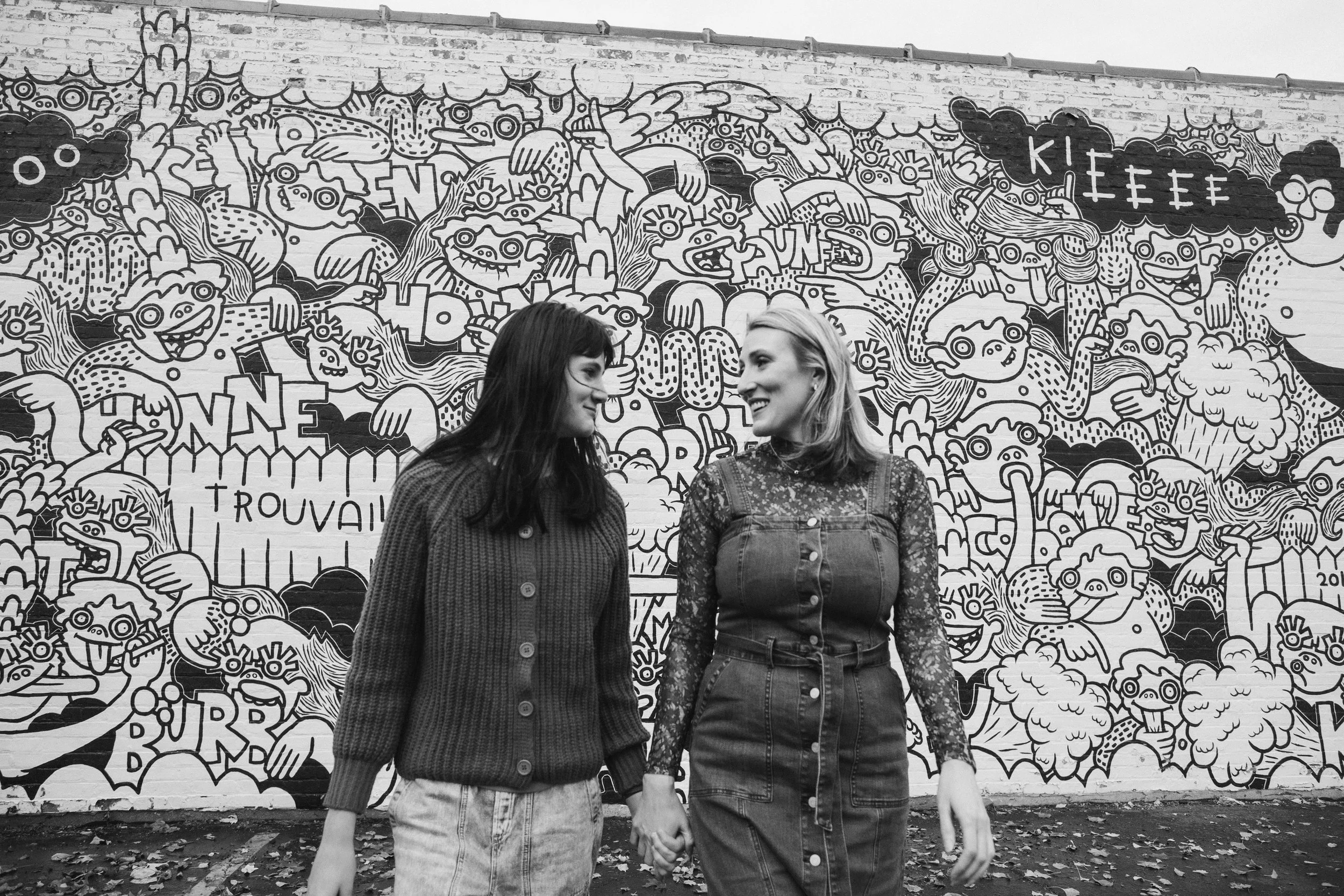 Two women holding hands and smiling at each other in front of a colorful graffiti mural on a brick wall, featuring cartoonish characters and text. Family photographer - Skokie IL