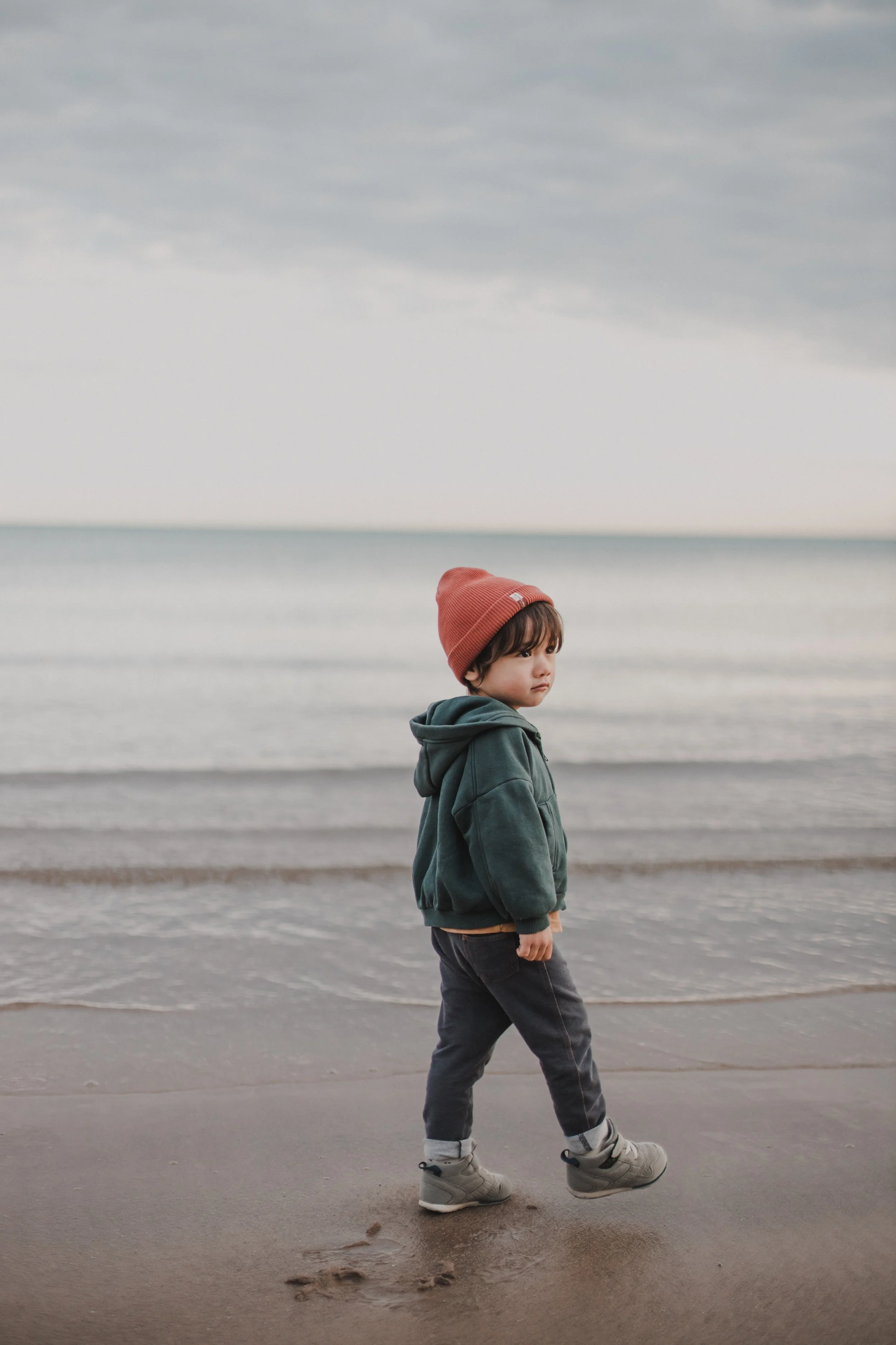 A young boy wearing a red beanie, grey hoodie, dark pants, and hiking shoes walking on the beach near the water with an overcast sky. Lake Michigan, Gillson Beach, Wilmette