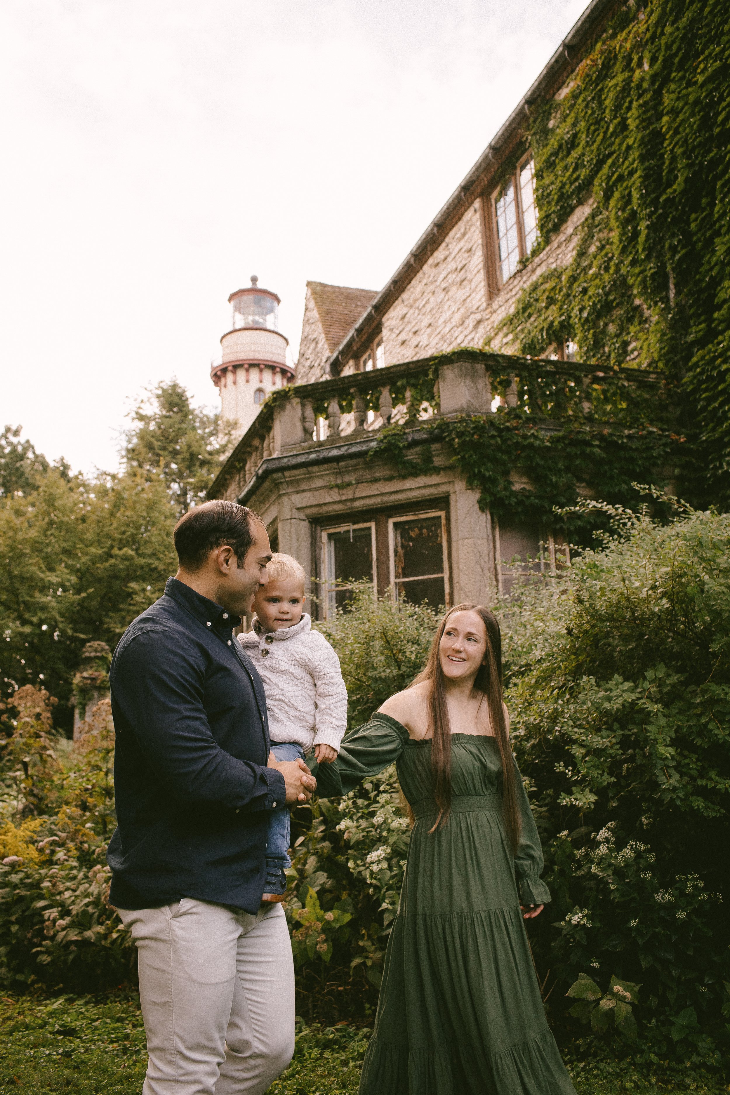 A family of three standing outdoors near a stone house with lush greenery and a lighthouse visible in the background. The man is holding a young girl, and the woman is smiling at them.