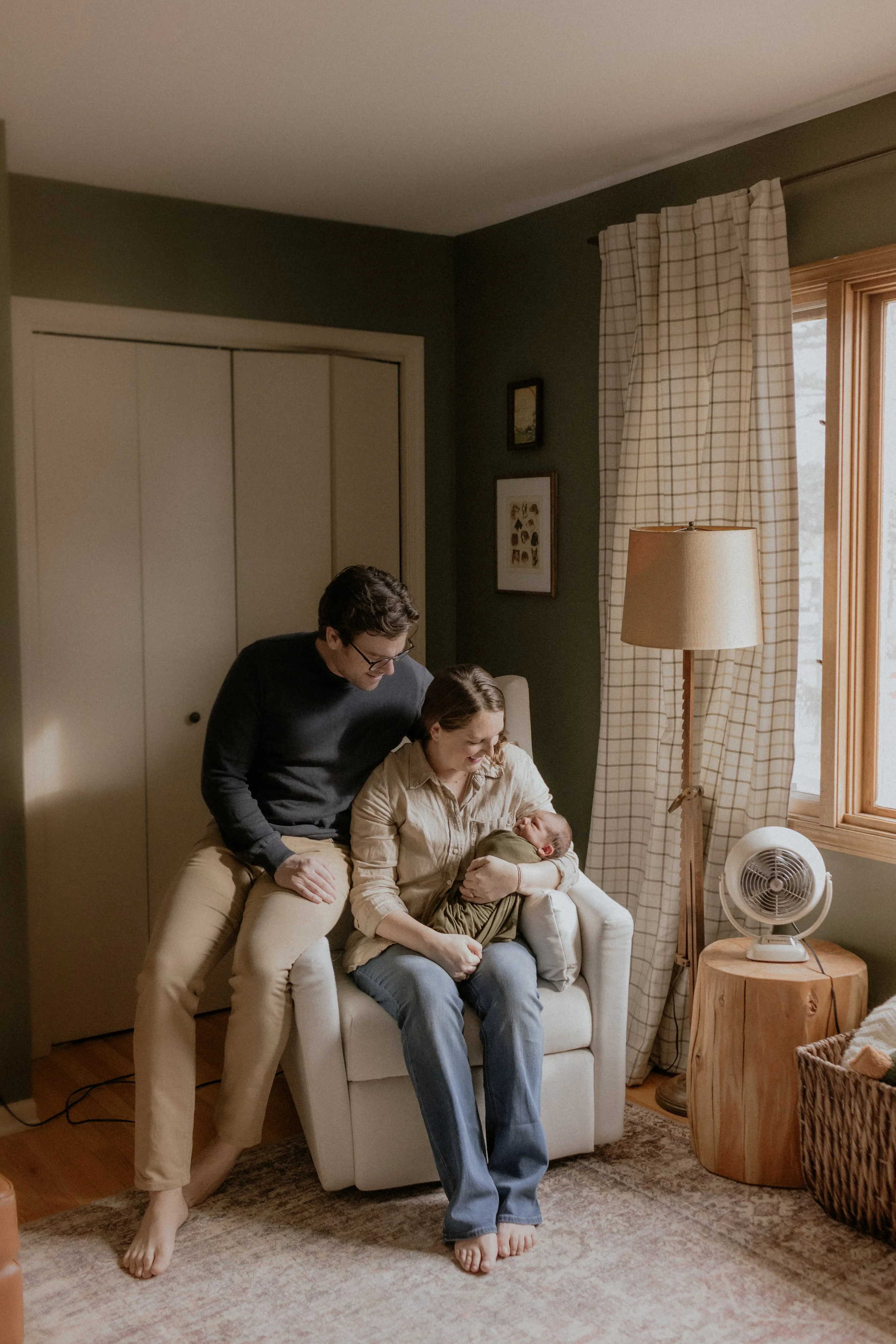 A couple sitting on a white armchair, looking at a newborn baby wrapped in a blanket. The woman is holding the baby, and the man is leaning over her, both smiling. Documentary newborn photography