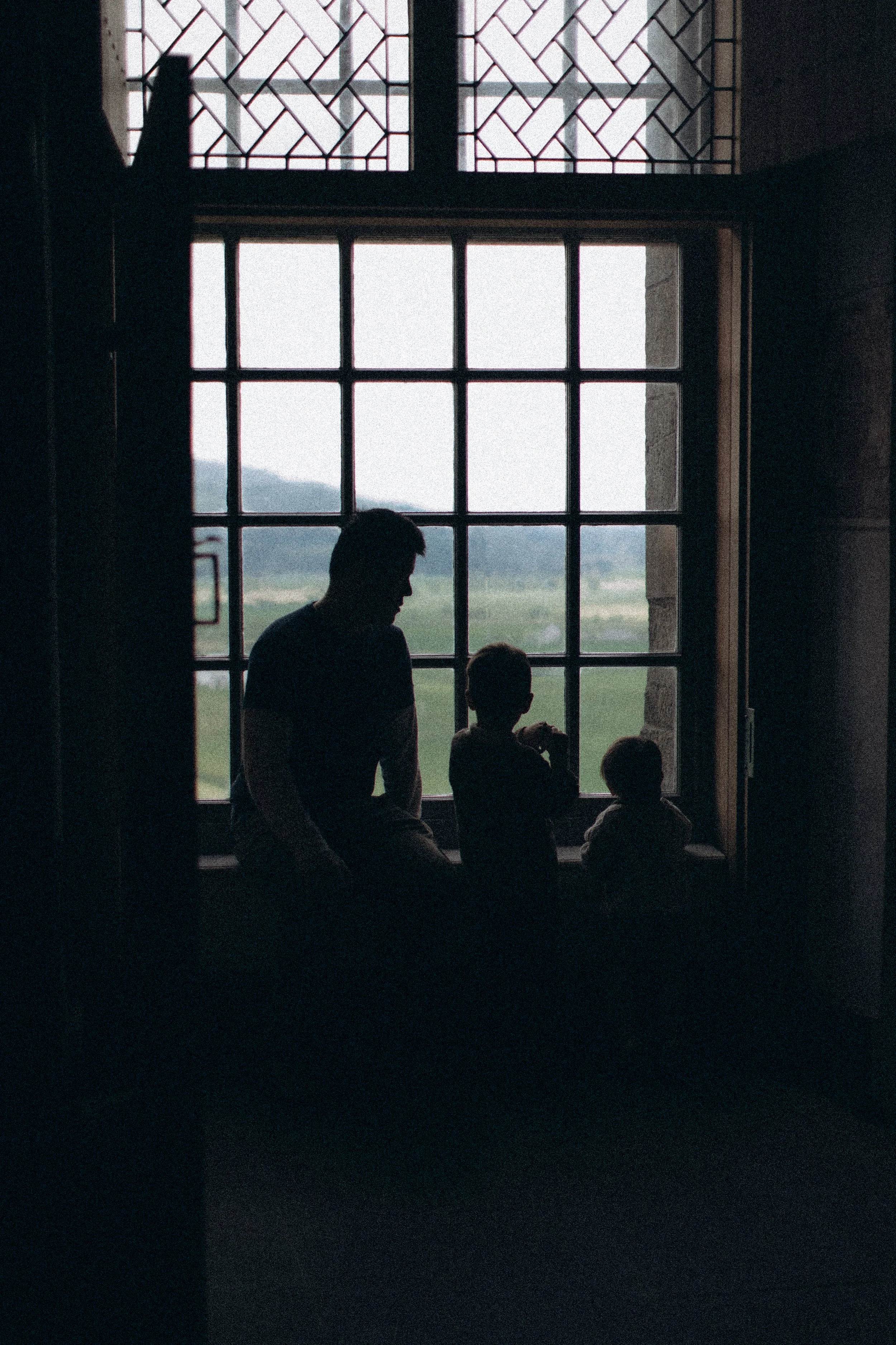Silhouettes of a man and two children sitting by a large window, with a scenic landscape visible outside, in dim interior lighting.