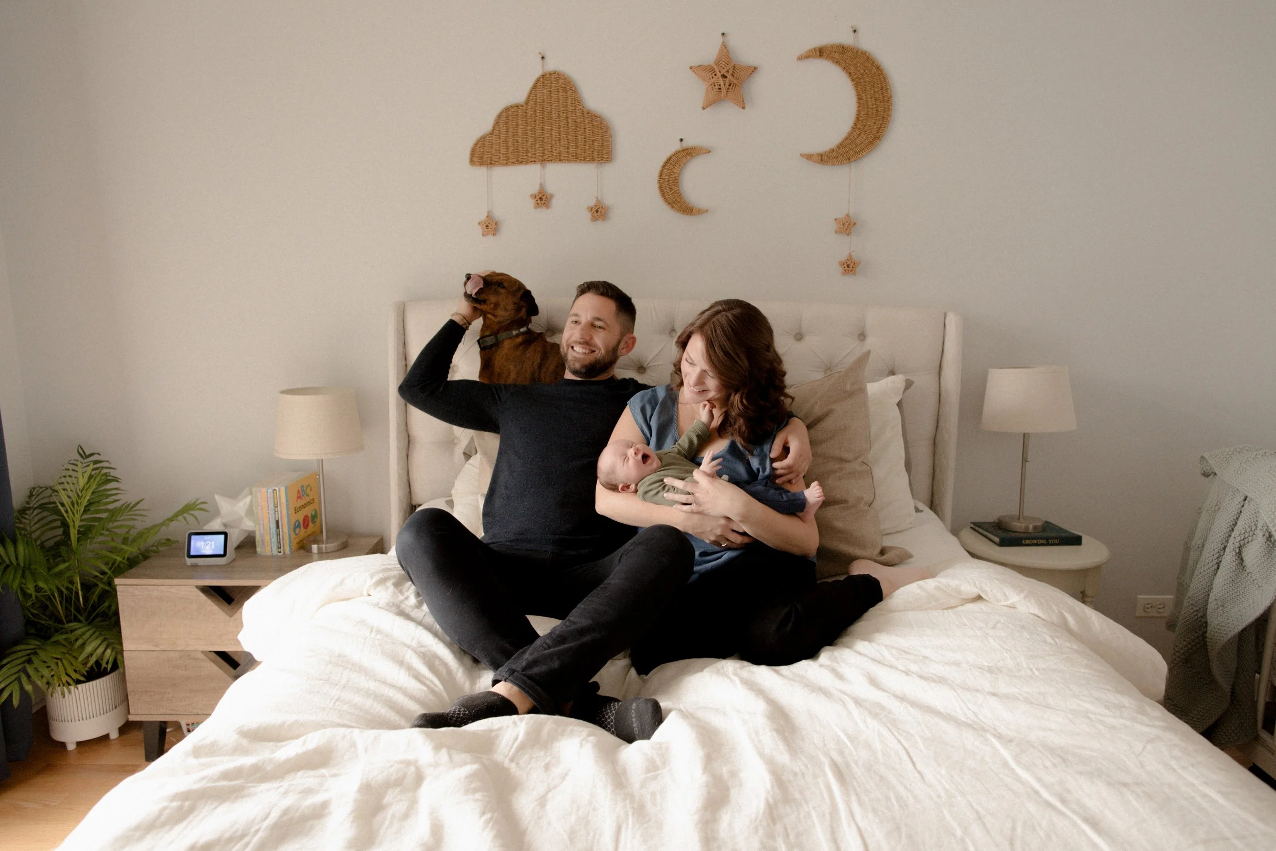 A happy family in a bedroom, with a man, woman, baby, and dog, sitting on a bed. The man is holding the dog, and the woman is holding the baby. Lifestyle newborn session shot in Chicago IL.