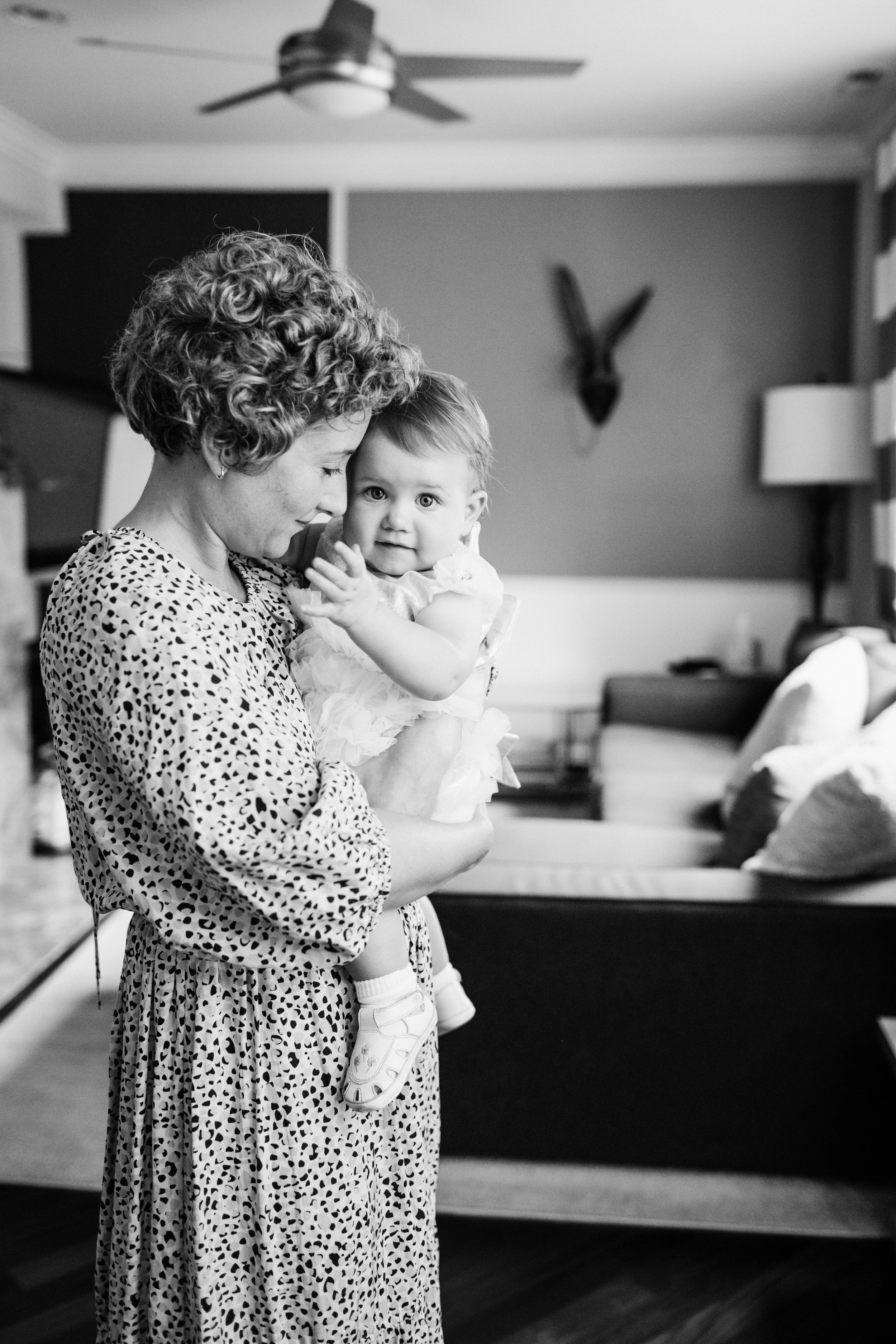 A woman with curly hair holding a baby girl in a living room. The woman is wearing a patterned dress, the baby is wearing a dress and shoes. The background shows a ceiling fan, a wall with a mounted head, and a couch with cushions.