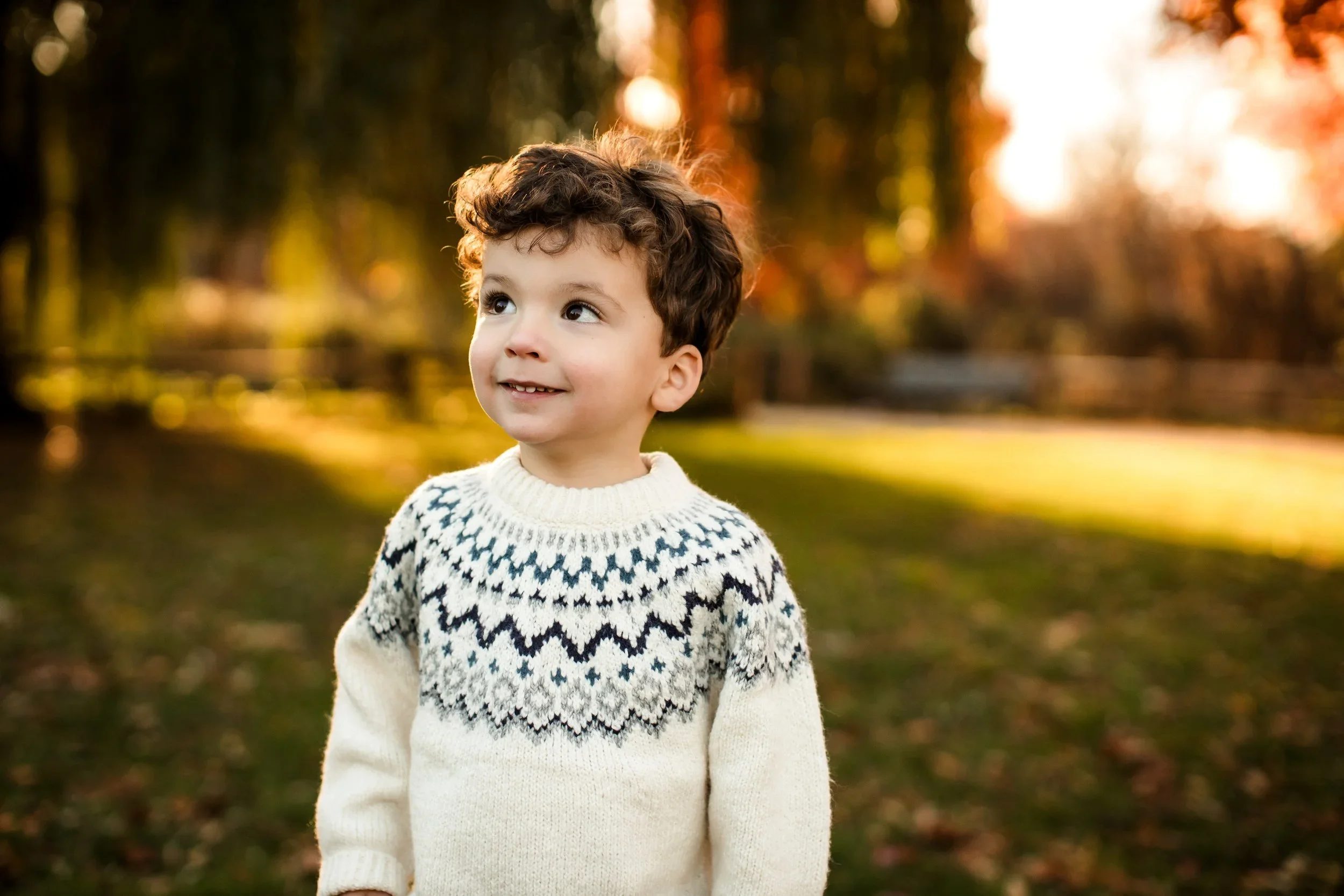 A young boy with curly brown hair smiling and looking up outdoors during autumn, wearing a white sweater with black and blue patterns. Blurred trees and fall foliage in the background. Chicago lifestyle family photography.  Shot at Winnemac Park.