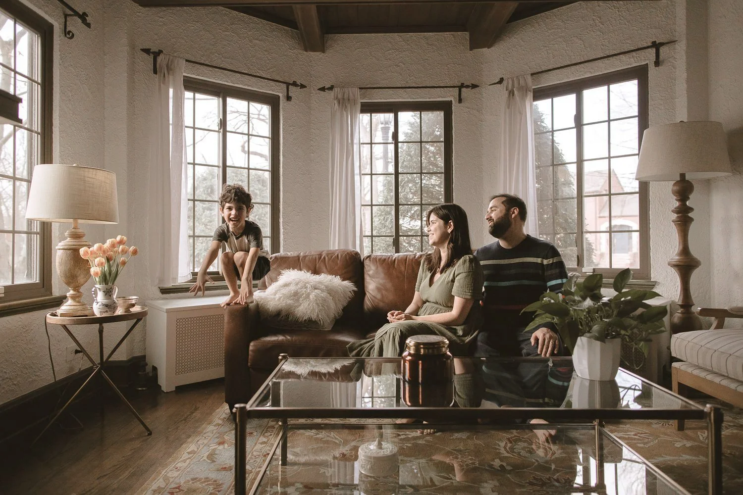 A boy sitting on a windowsill laughing, with a woman and a man sitting on a leather sofa in a living room, sunlight coming through large windows, houseplants and lamps. Lifestyle Family Photography Session Evanston, IL.