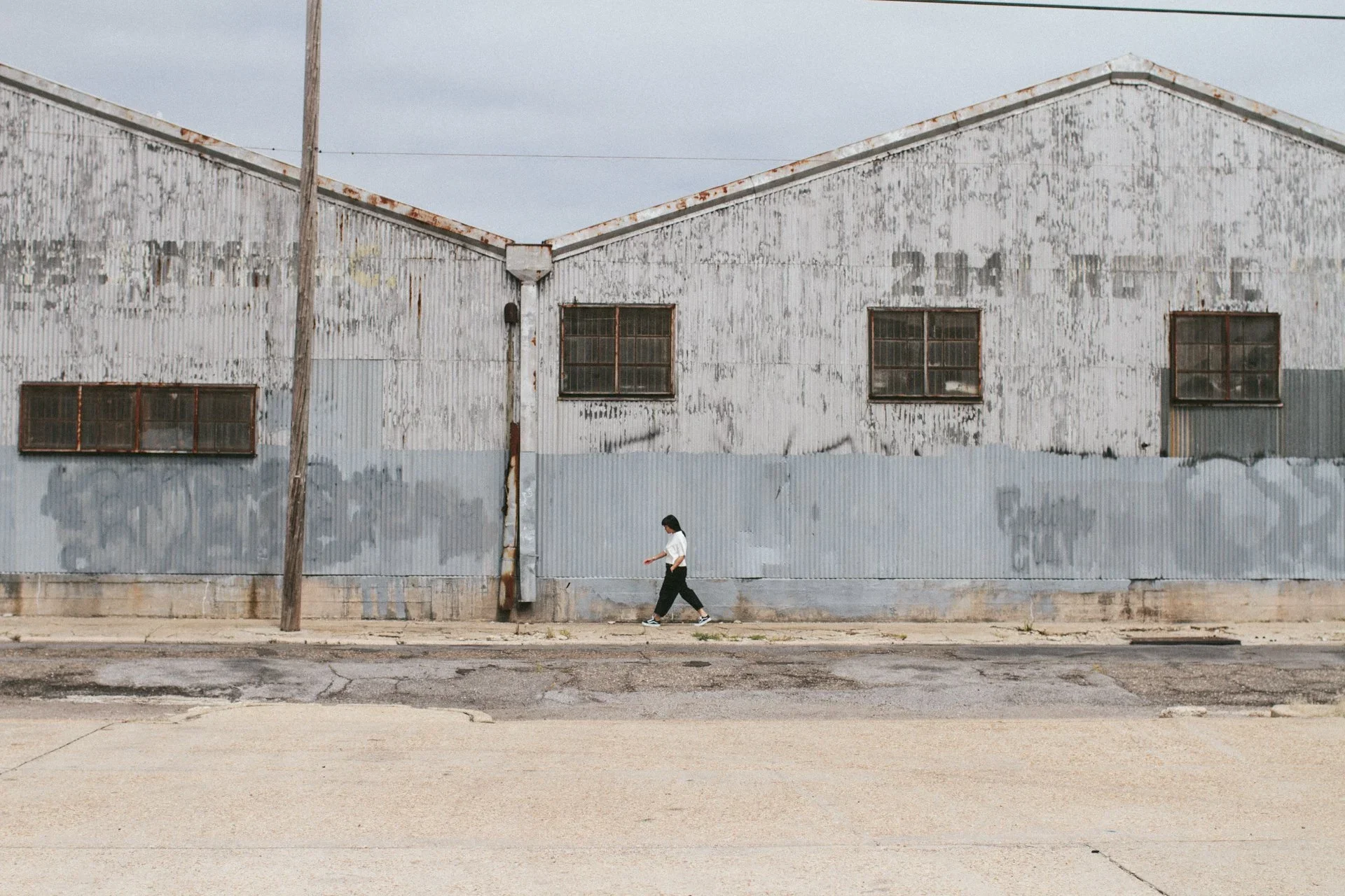 A woman walking past an old, weathered industrial building with rusted metal siding and broken windows.