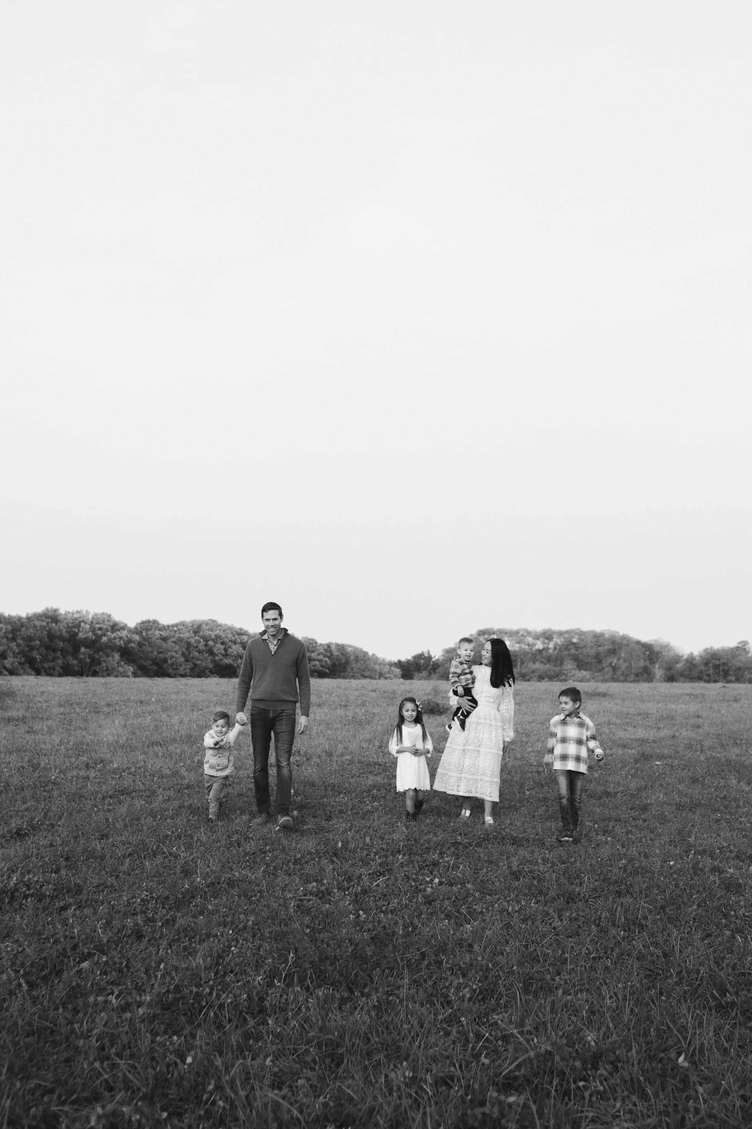 A family of six walking in a grassy field on a cloudy day, holding hands and carrying a young child.