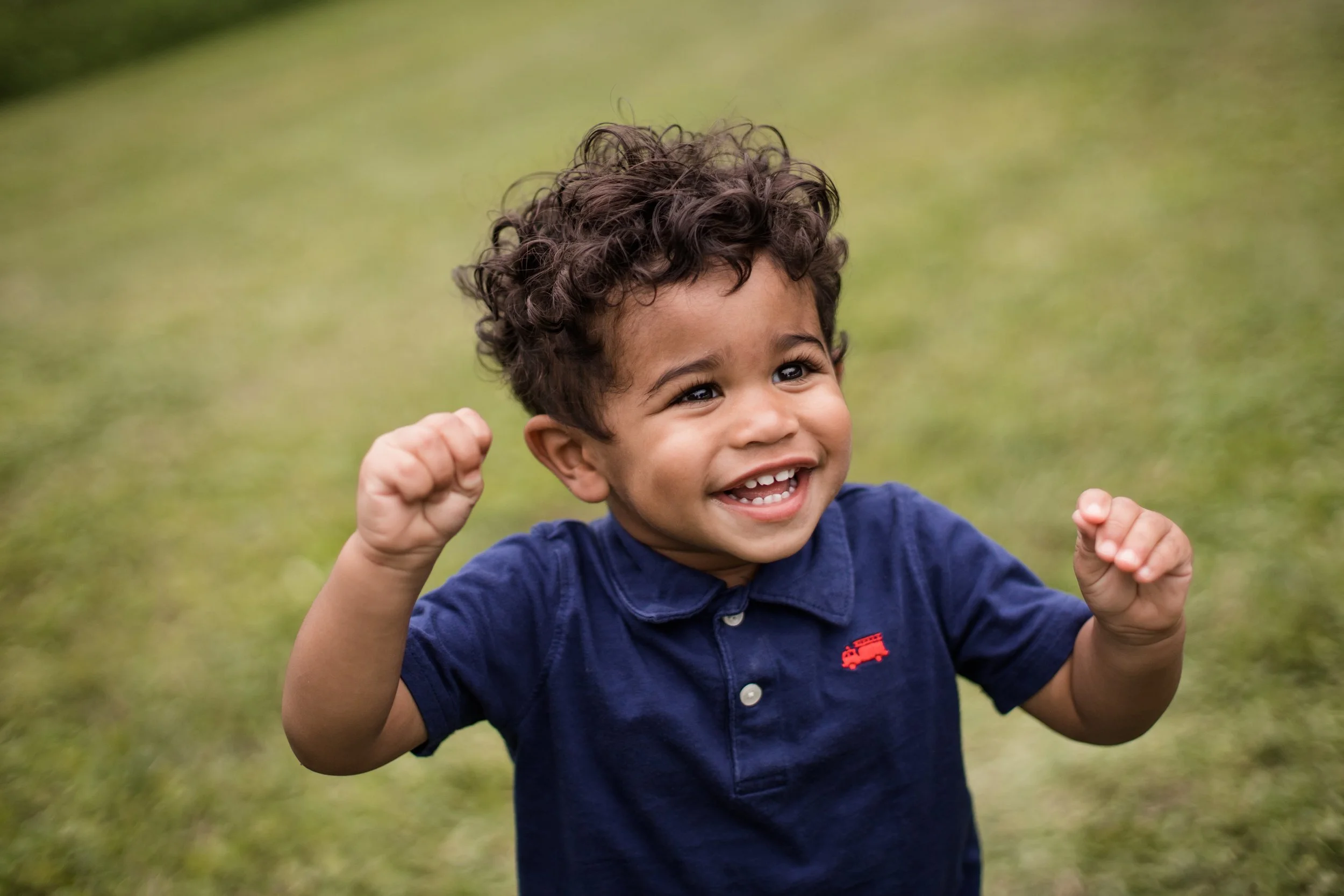 A young child with curly hair, smiling and raising fists in excitement, standing outdoors on green grass.  Documentary Family Photographer in Evanston and Chicago IL.
