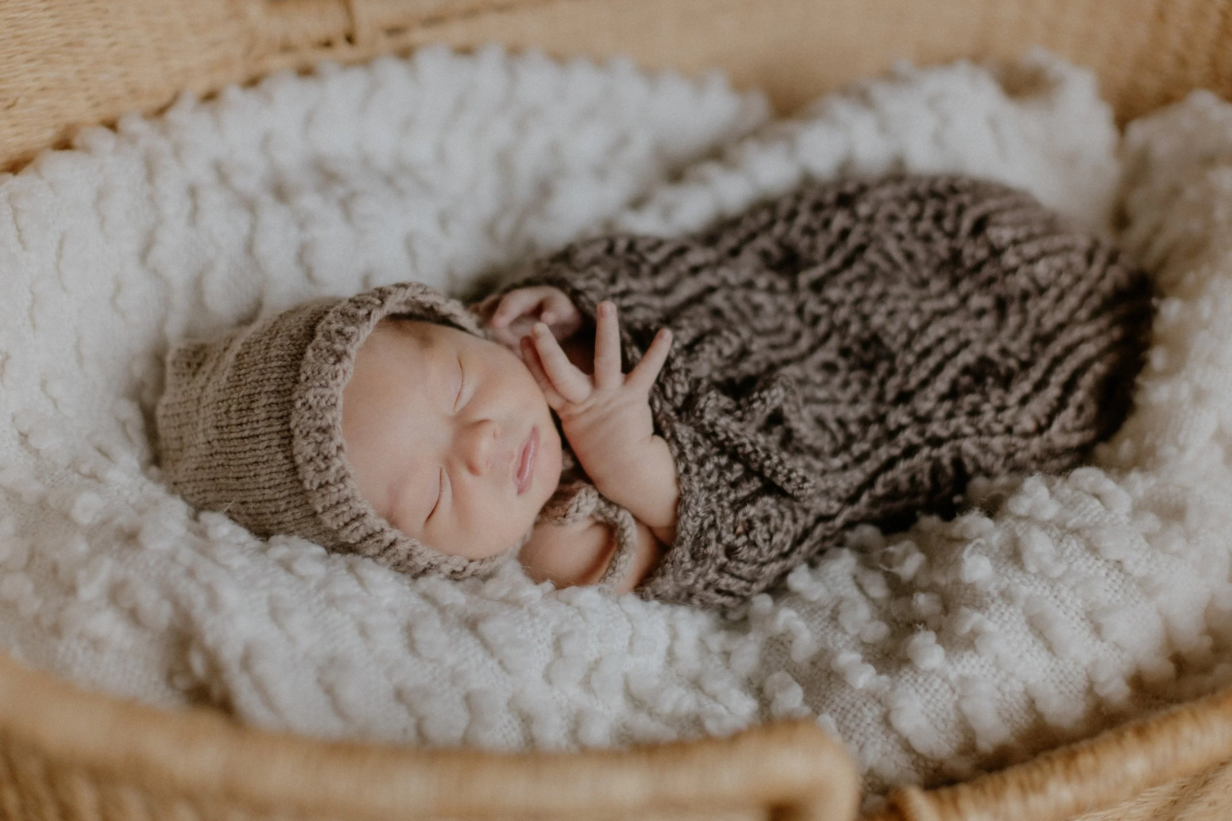 A sleeping newborn baby wearing a knitted hat and outfit, lying on a soft white blanket in a woven bassinet.