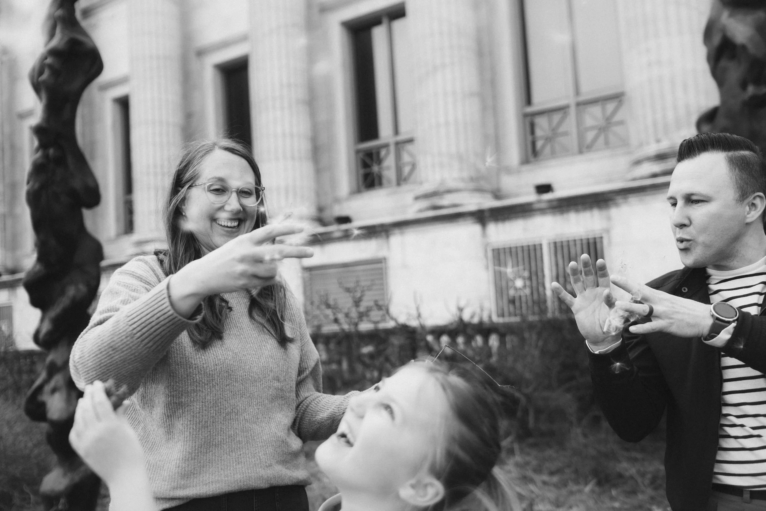 family outdoors, smiling and engaging with a large soap bubble. The woman on the left wears glasses and a sweater, while the man on the right wears a striped shirt and a jacket. A building with windows and balconies is in the background.
