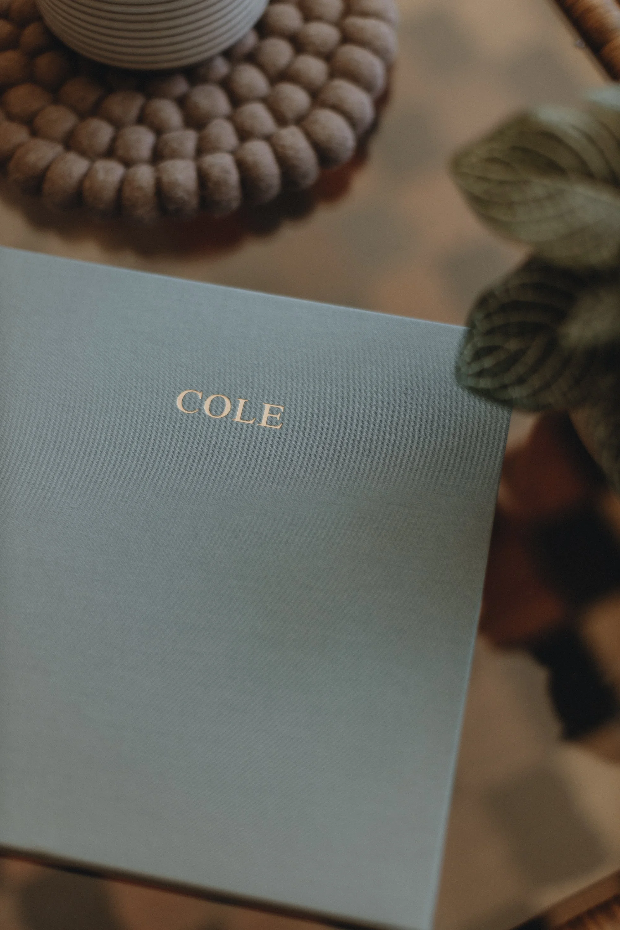 A light blue card with the name 'COLE' written on it. The card is placed on a surface near a woven placemat with a bowl on top and some decorative dried leaves nearby.