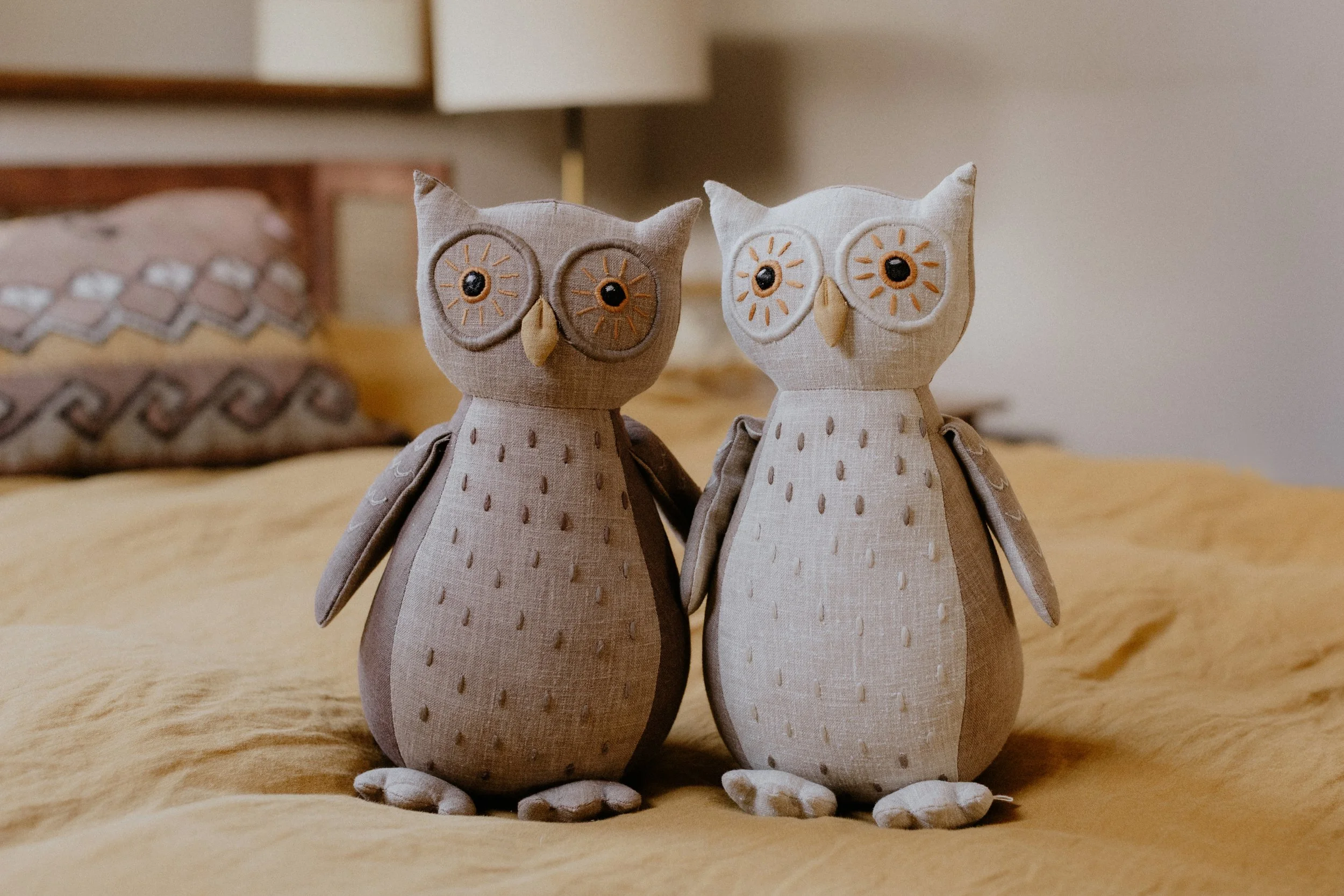 Two fabric owl plush toys sitting on a bed with beige sheets and a decorative pillow in the background. Product Photography Shoot Evanston, IL.