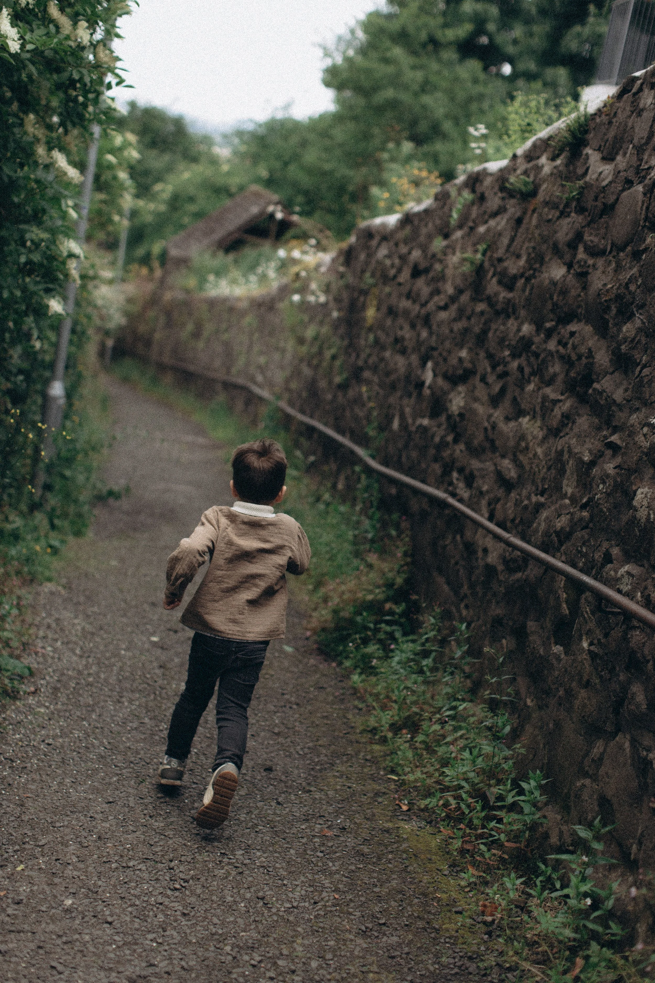 A young boy running along a narrow dirt path surrounded by lush green bushes and trees, with a stone wall on his right side.