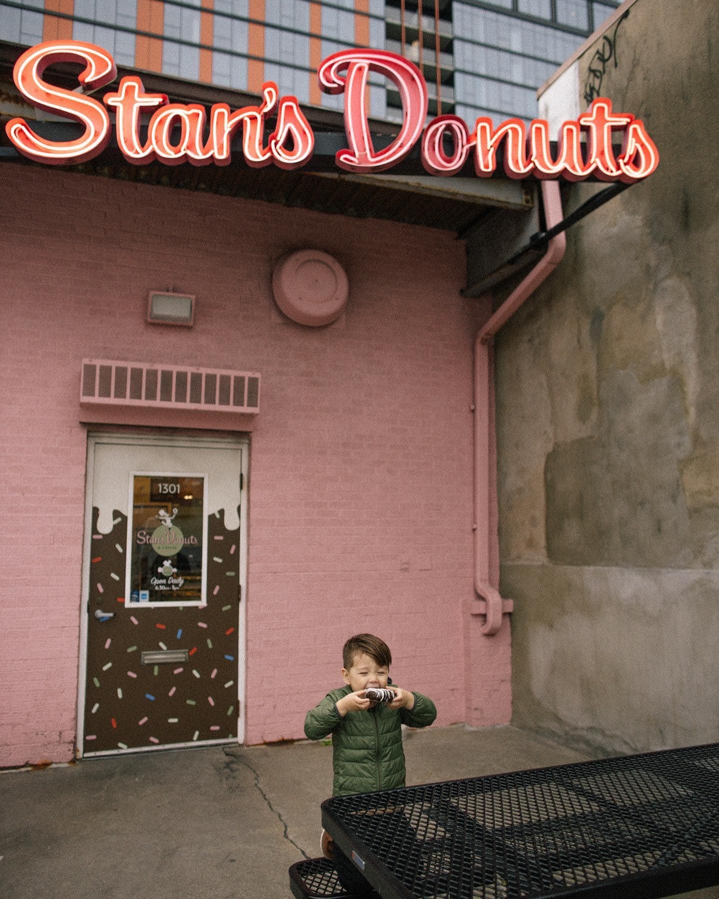 A young boy in a green jacket taking a photo with a camera outside a pink donut shop called "Stan's Donuts" with a pink and brown donut-pattern door.