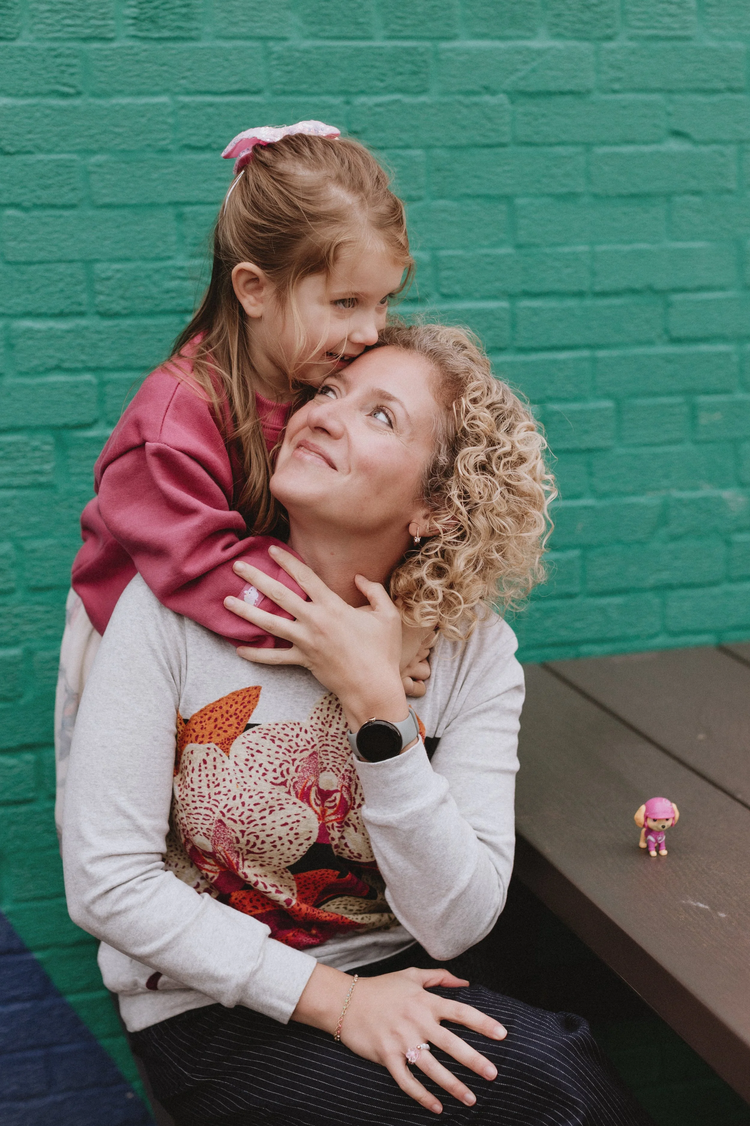 A woman and a young girl sharing a tender moment, with the girl on the woman's shoulders. The girl is leaning in close to the woman, who is looking up at her affectionately. They are seated in front of a green brick wall, and there is a small toy on 