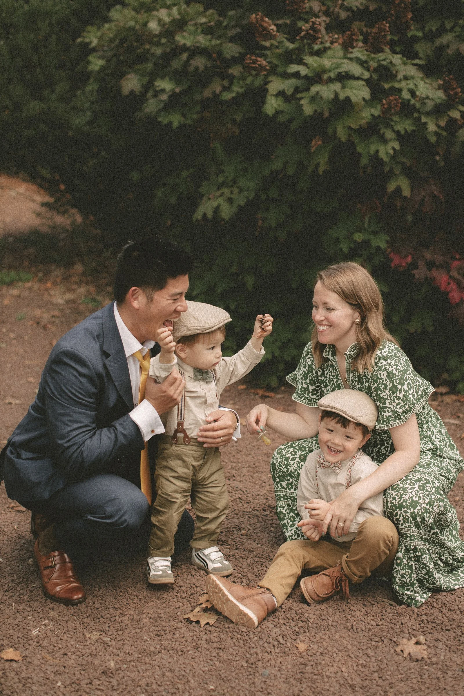 A happy family of four, including two children and two adults, enjoying time outdoors on a dirt path surrounded by greenery and foliage. The children are wearing beige hats and casual clothing, and the adults are smiling and interacting with the kids.