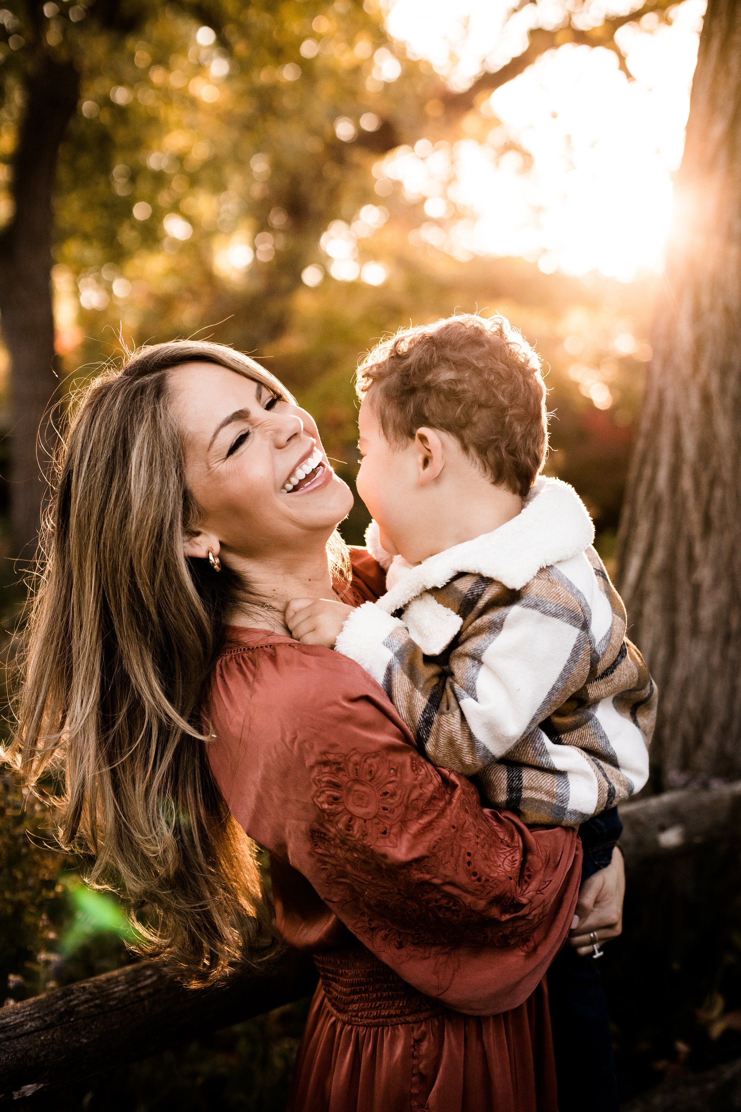 A woman and young boy sharing laughter outdoors in a park during sunset with fall foliage in the background.