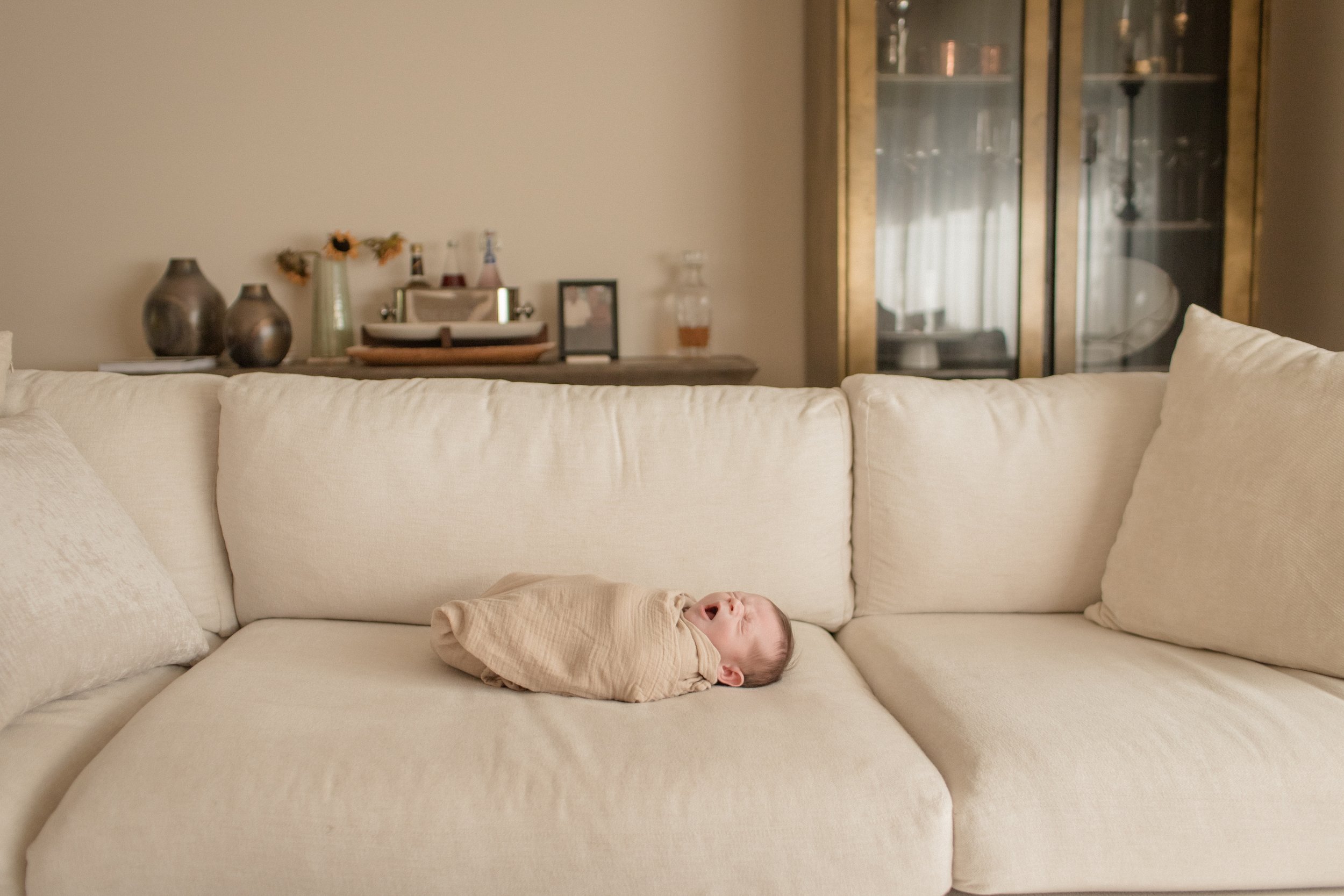 A yawning baby wrapped in a beige blanket lying on a cream-colored sofa in a living room. Lifestyle newborn session shot in Evanston IL.
