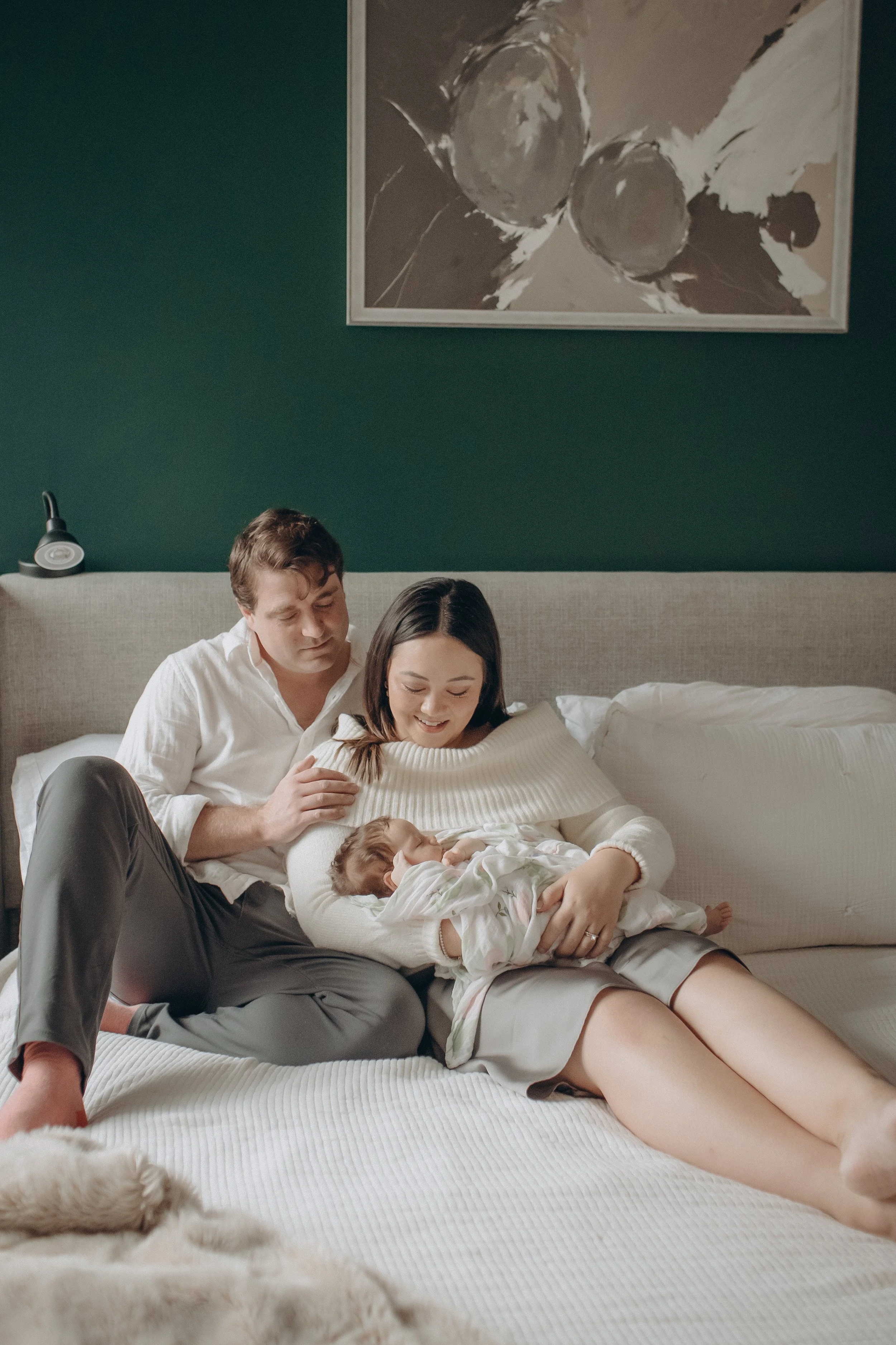 A family of three, including a mother, father, and their newborn baby, sitting on a bed together, smiling and bonding. Lifestyle newborn session shot in Chicago IL.