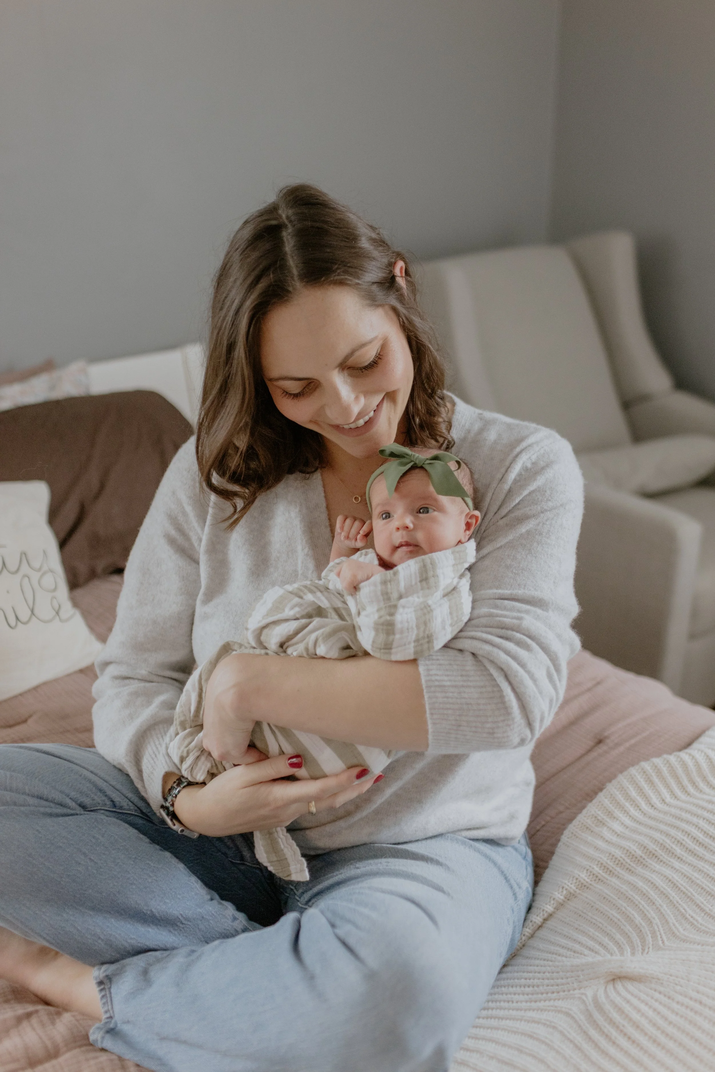A woman sitting cross-legged on a bed, smiling and holding a baby girl with a green bow headband, in a cozy room with neutral-colored furniture. Lifestyle newborn session shot in Evanston IL.