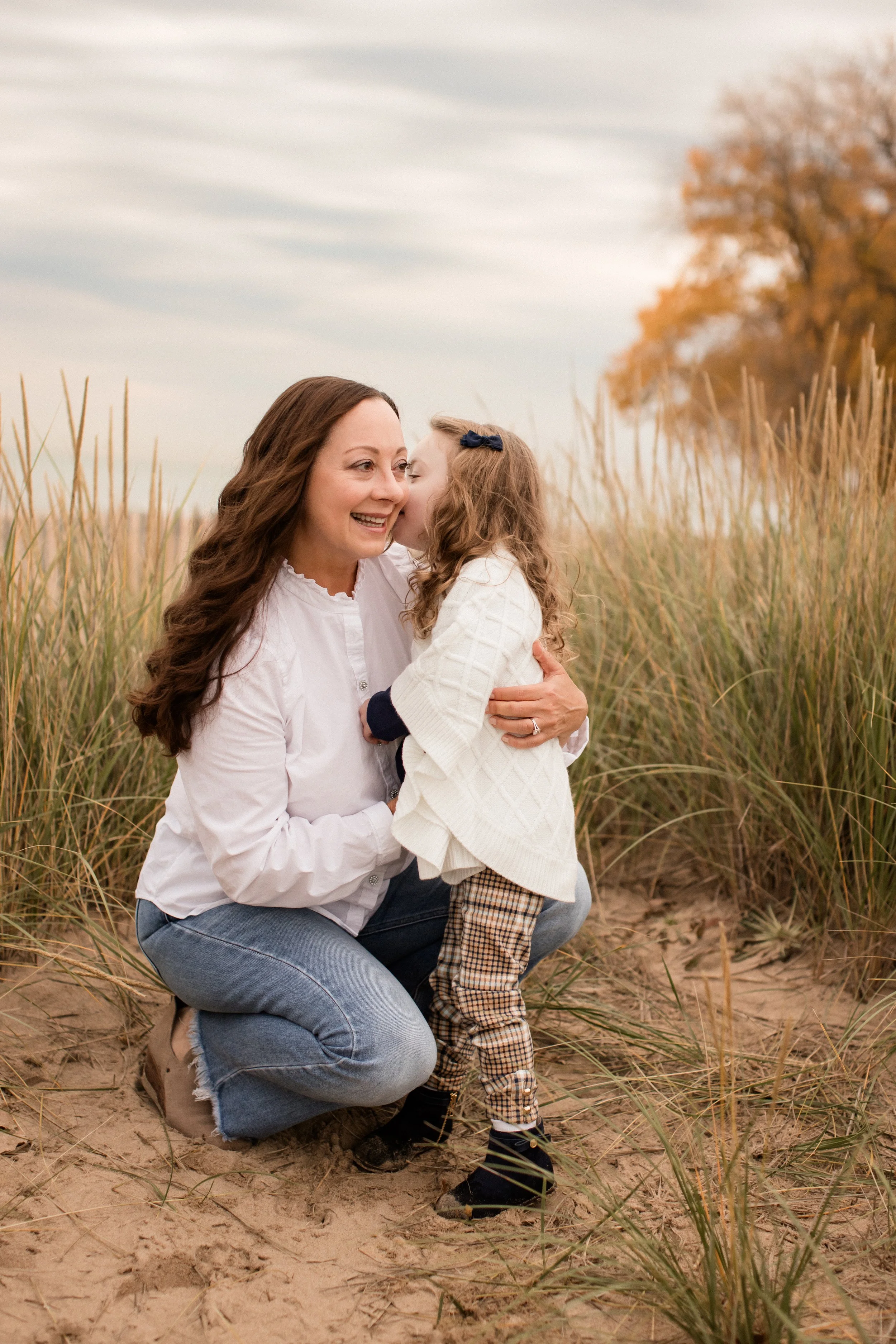 A woman kneeling in the sand, hugging a young girl and sharing a kiss on her cheek, with tall grass and trees in the background during the fall.