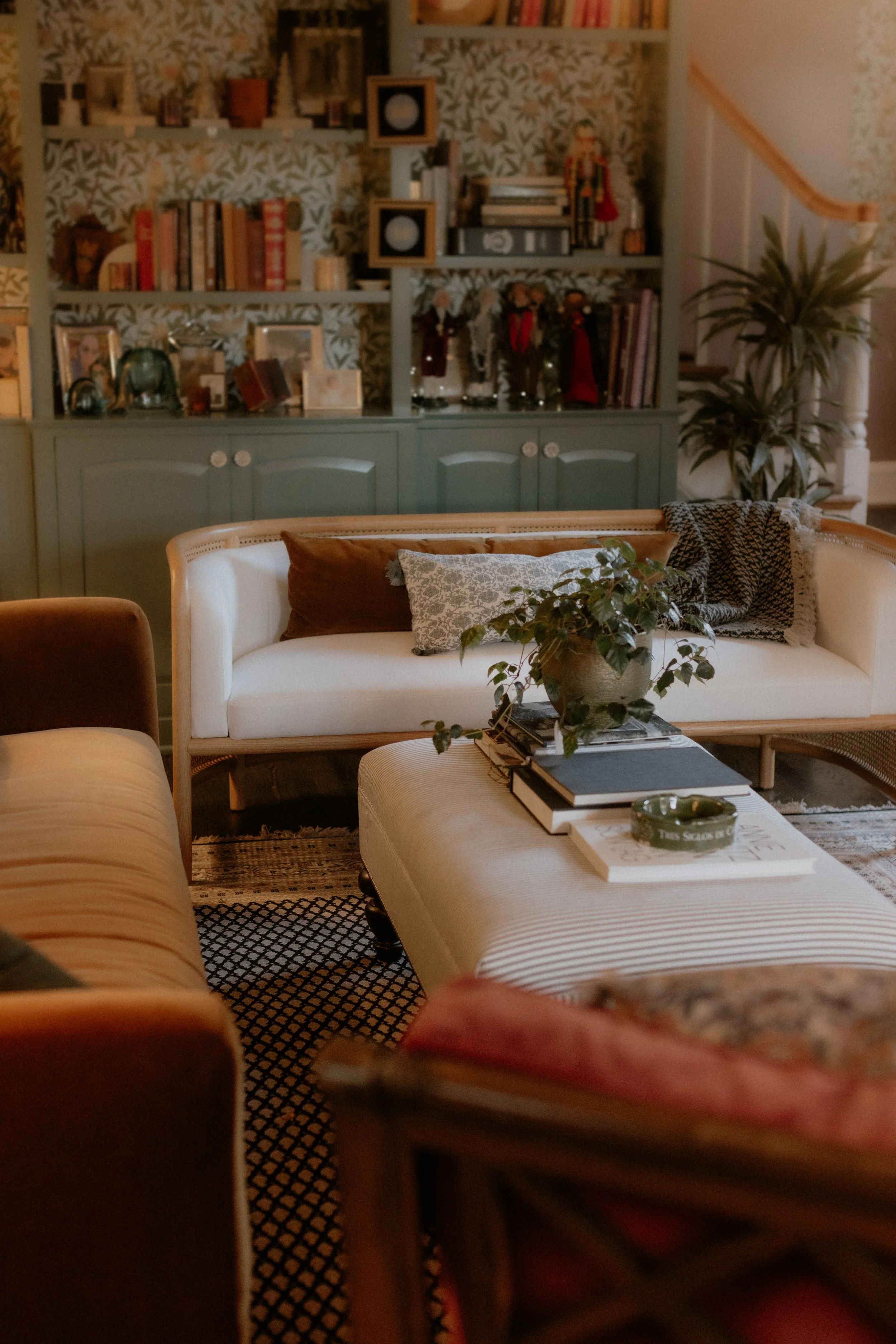 A cozy living room with a white sofa, brown and patterned pillows, a plant on a tray, books, and decorative items on shelves against a patterned wallpaper background.  Interior Design Photography Chicago IL, Erin Drewitz Photography