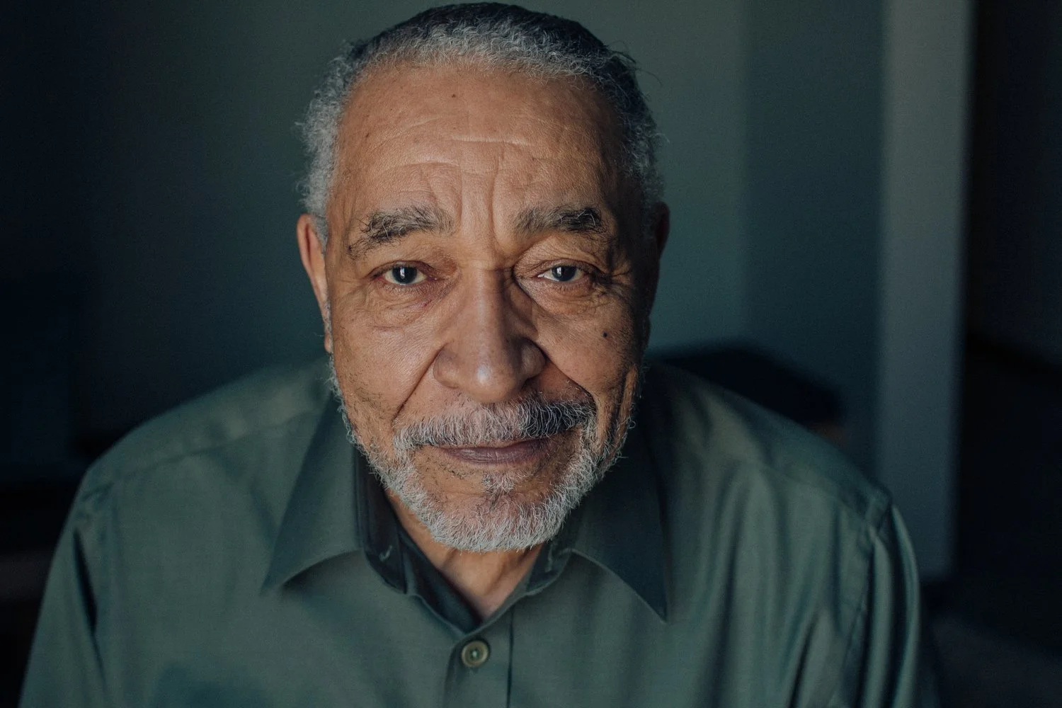 Close-up of an elderly man with gray hair, beard, and mustache, wearing a dark green shirt, looking at the camera with a gentle expression.
