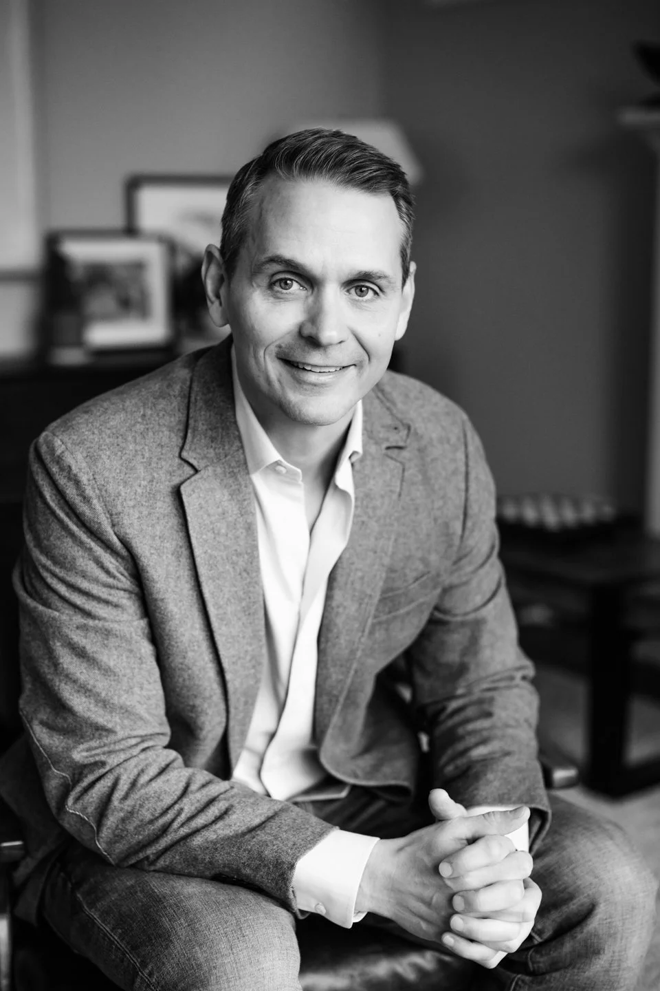 A black-and-white photo of a man in a suit jacket and white shirt, sitting with hands clasped, smiling, in an office setting.