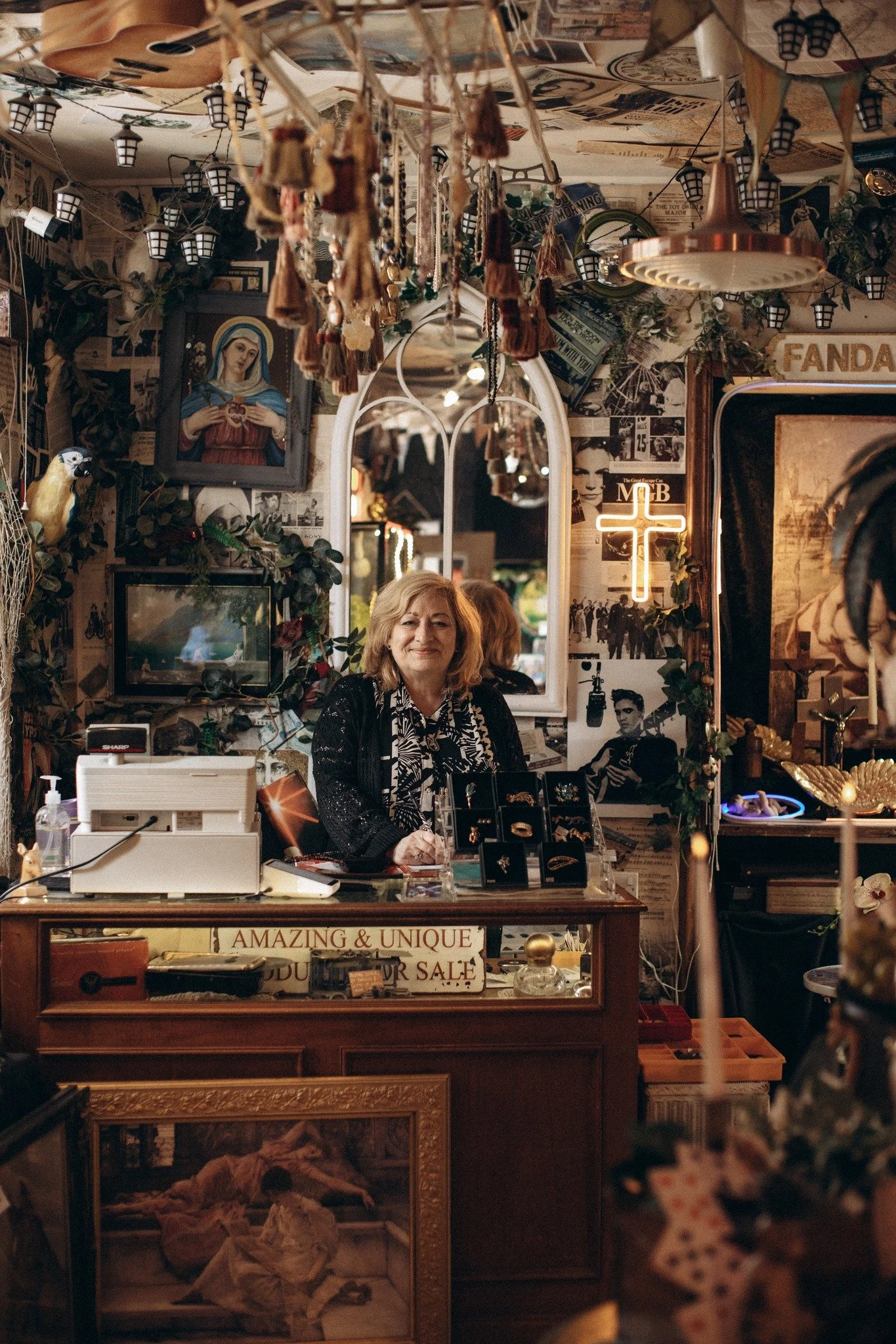 A woman standing behind a sale counter in a vintage store, decorated with various eclectic items, artwork, and a neon cross sign on the wall.