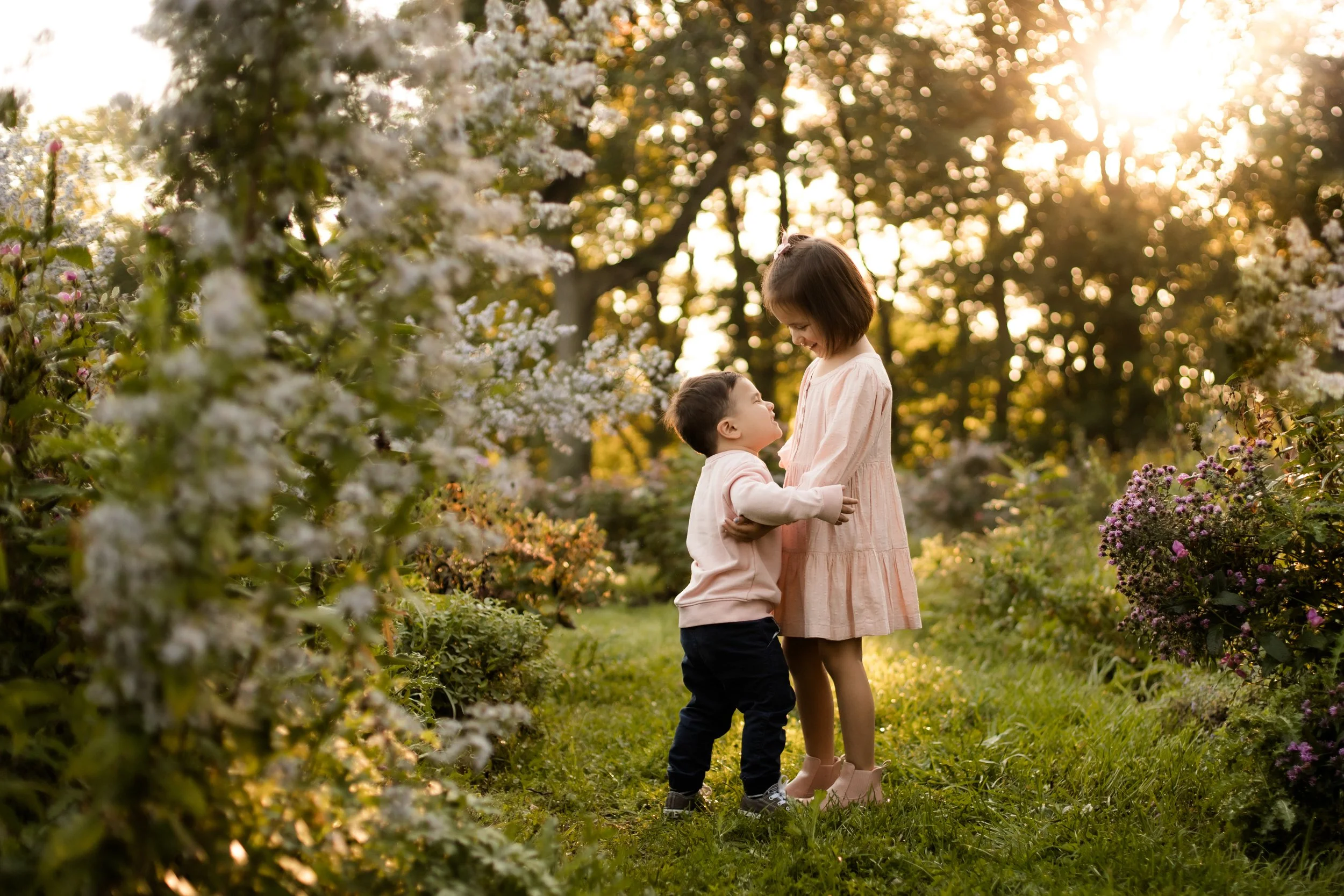 A young boy and girl standing close together in a garden with flowering bushes during sunset, with the boy looking up at the girl and holding her arms, and the girl smiling down at him.